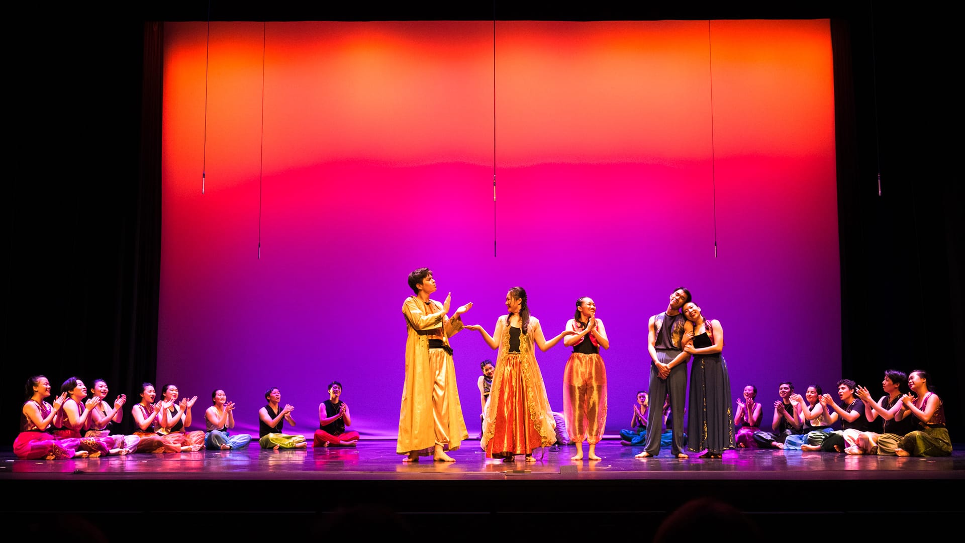 Performers in colorful costumes on a stage with a vibrant orange-to-purple gradient backdrop, surrounded by seated cast members.