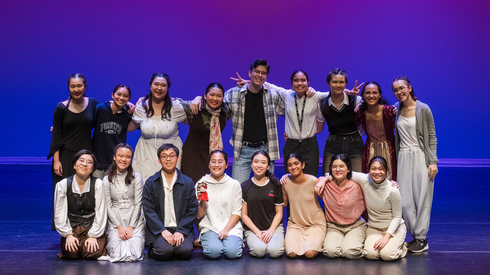 A group of about 15 smiling young people posing together on a stage with purple and blue lighting.