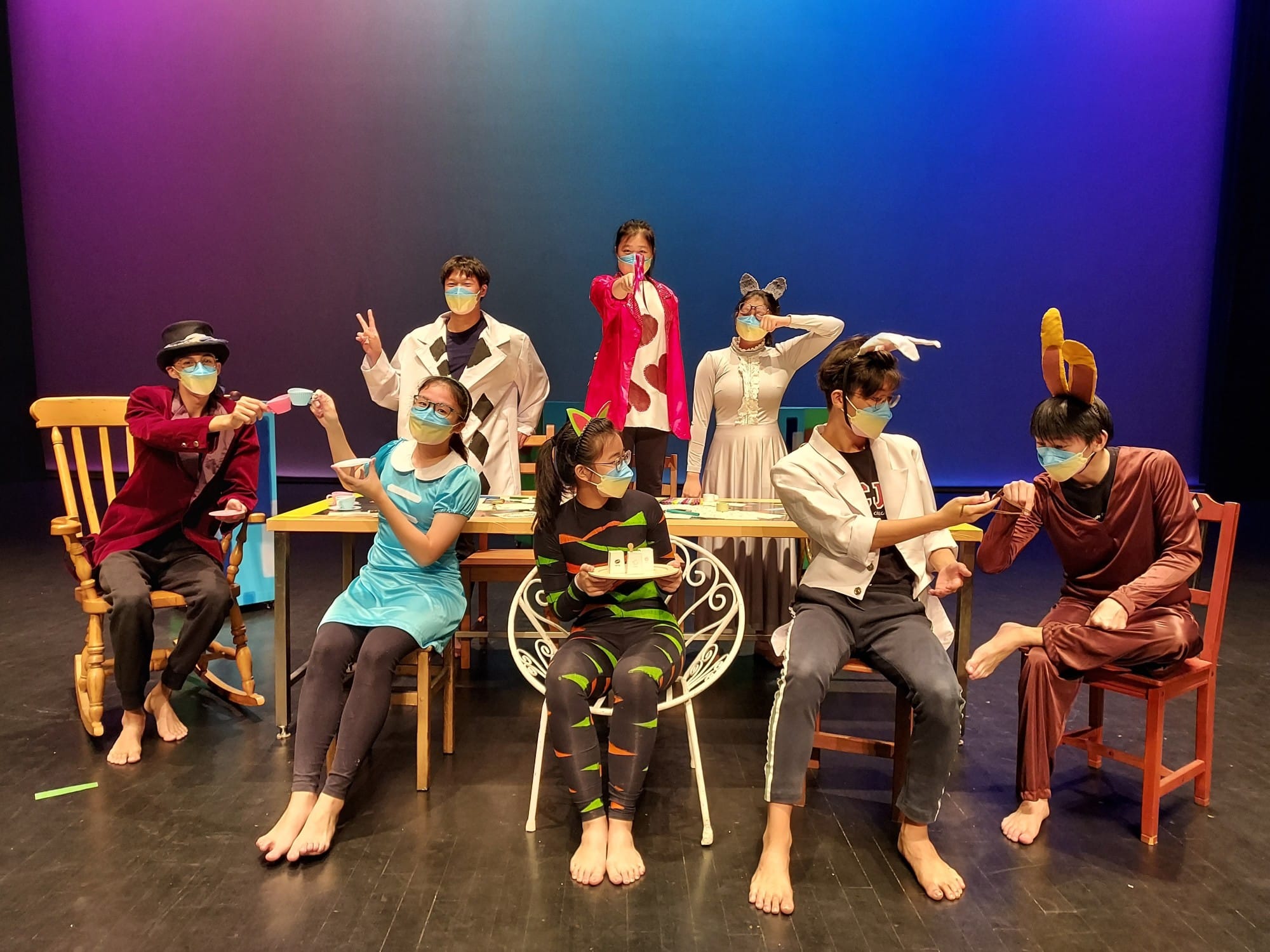 Eight masked performers in colorful Alice in Wonderland costumes pose on a stage with chairs and a table holding teacups.