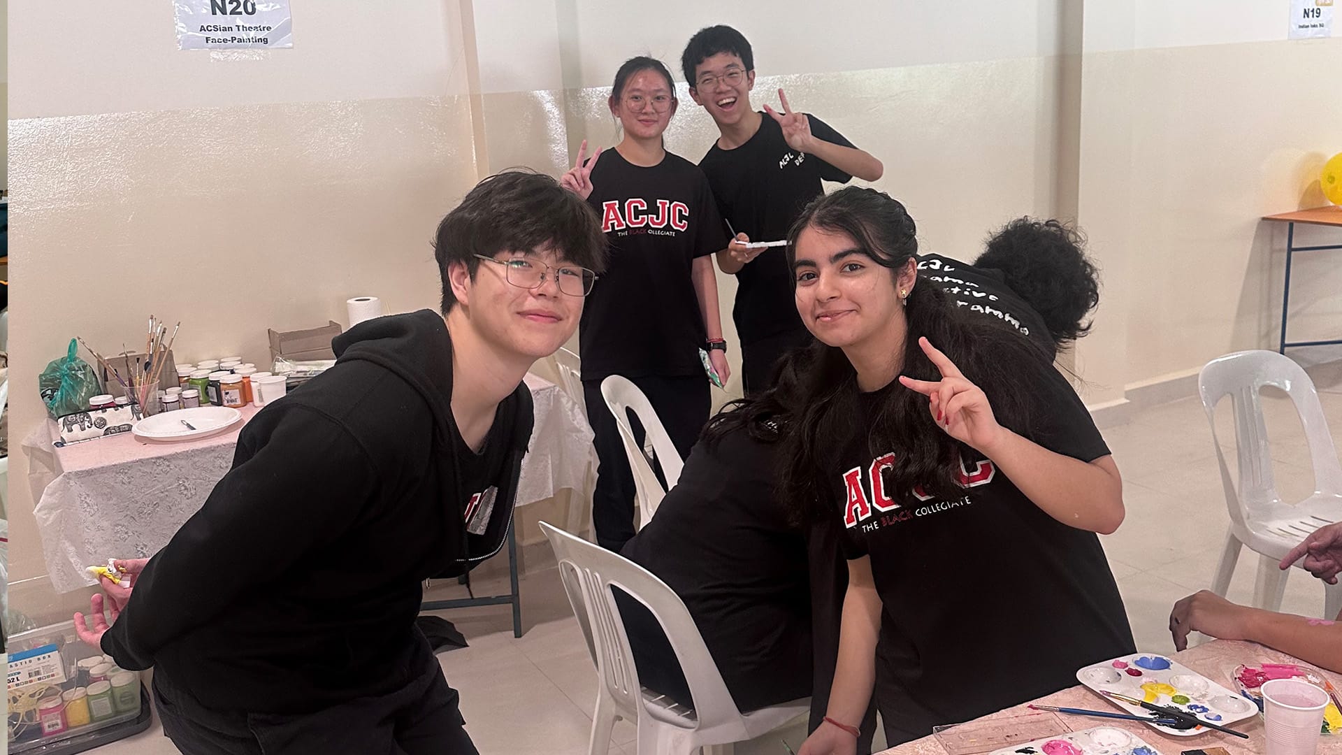 Four smiling students in black ACJC shirts pose with peace signs at a face-painting booth with paint supplies on a table.