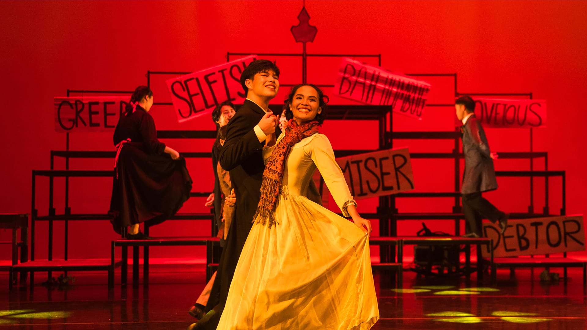 Two smiling performers dance on a red-lit stage with signs reading "Selfish," "Miser," "Greedy," and "Debtor" behind them.
