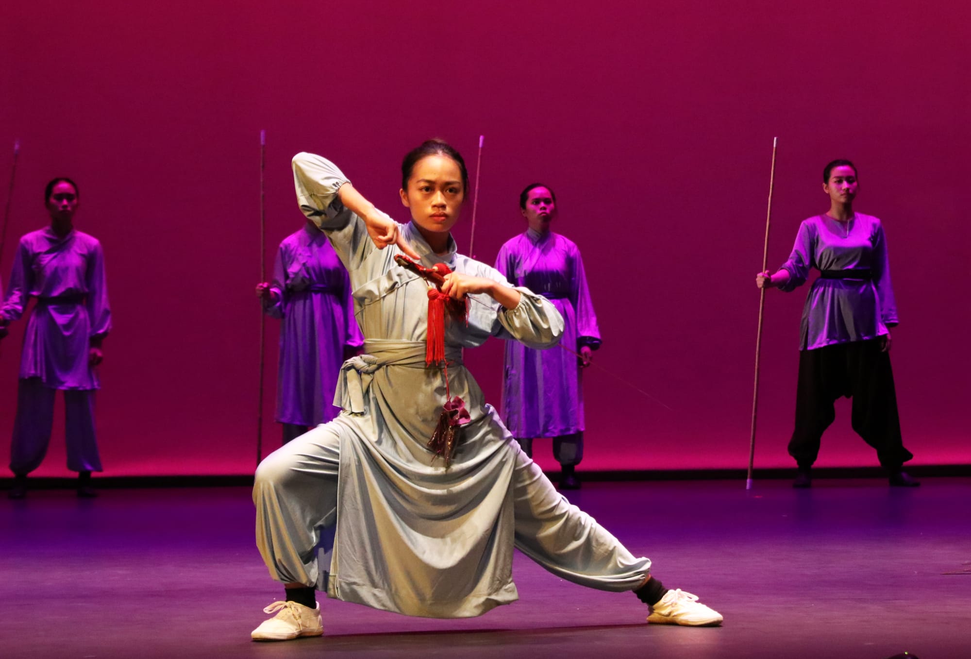 A woman in gray wushu attire holds a sword in a low stance on stage, with four performers holding staffs behind her.