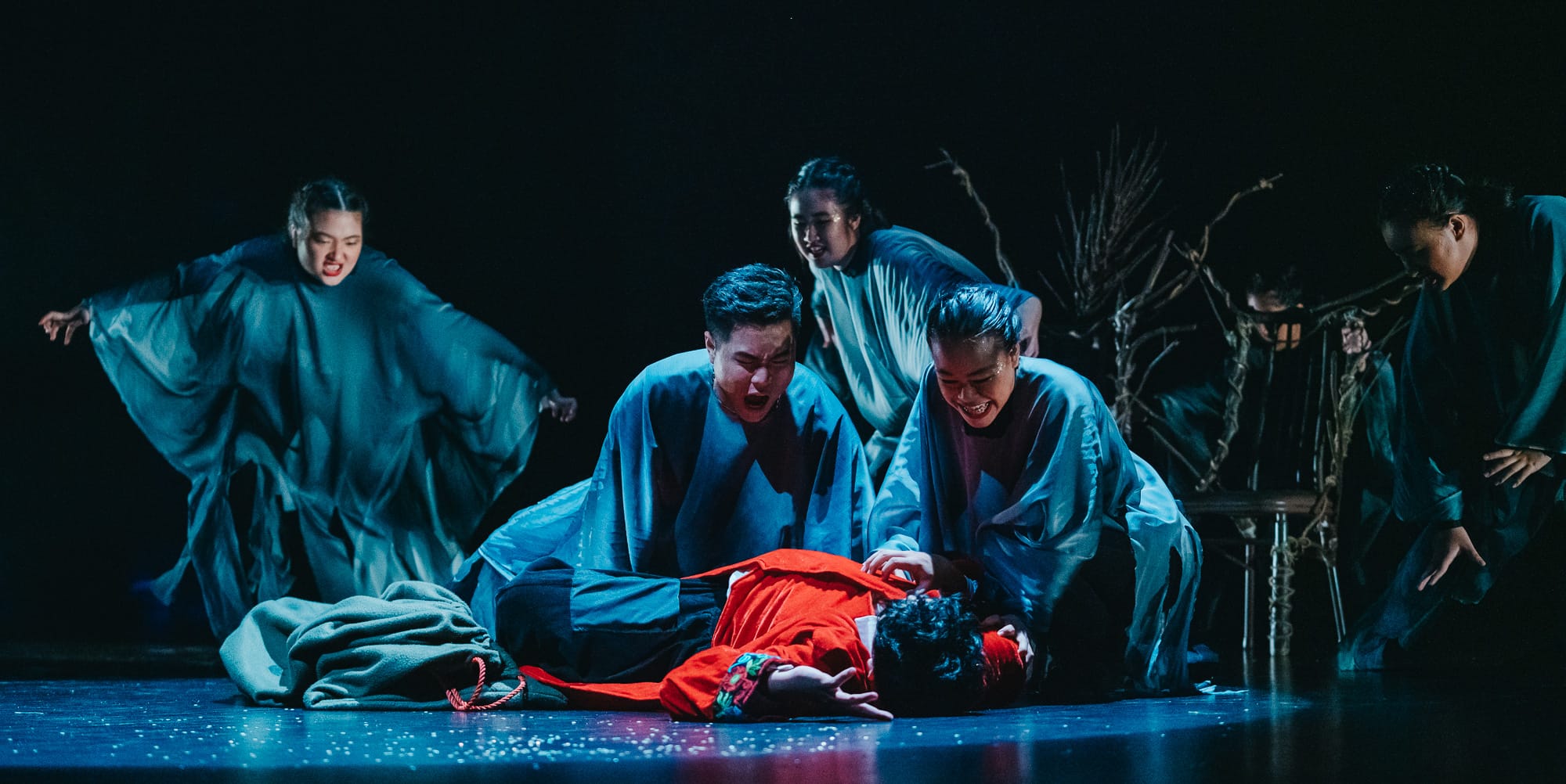 Performers in blue robes surround a figure in red lying on a dark stage in an intense theatrical scene.