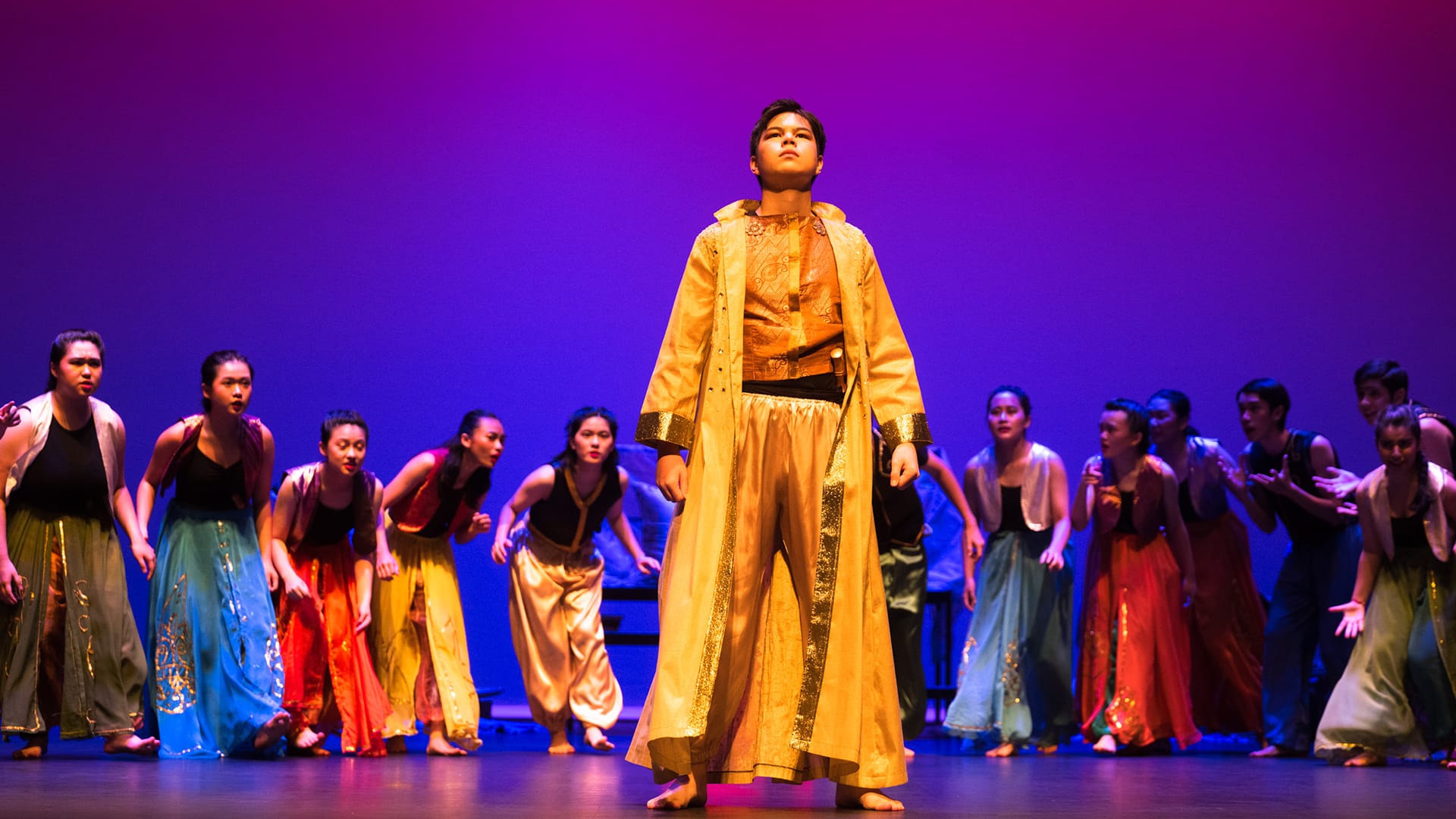 A performer in a gold costume stands center stage while dancers in colorful costumes pose behind under purple lighting.