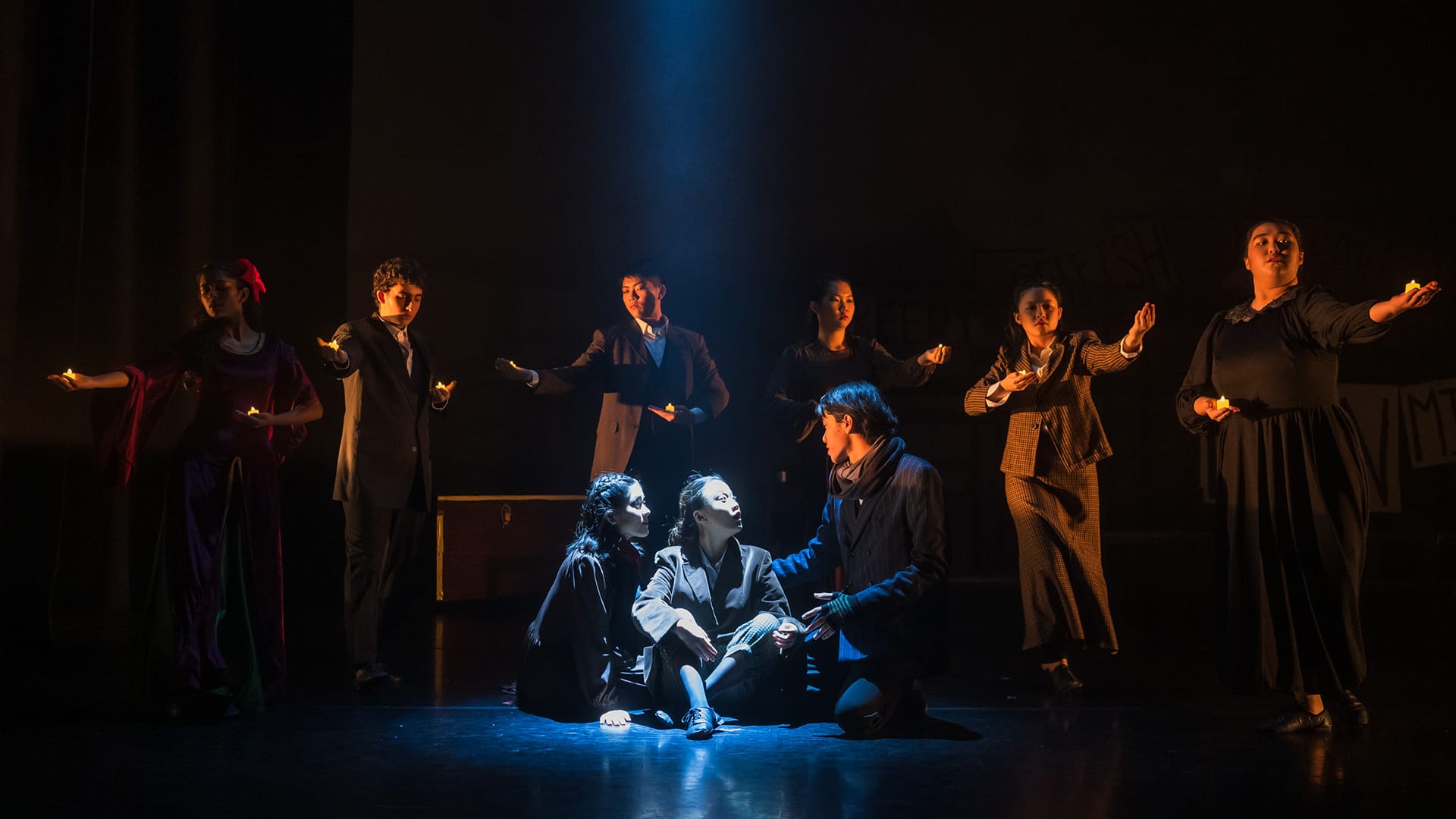 Theater performers on a dark stage, two kneeling in a blue spotlight while others stand holding candles with arms outstretched.