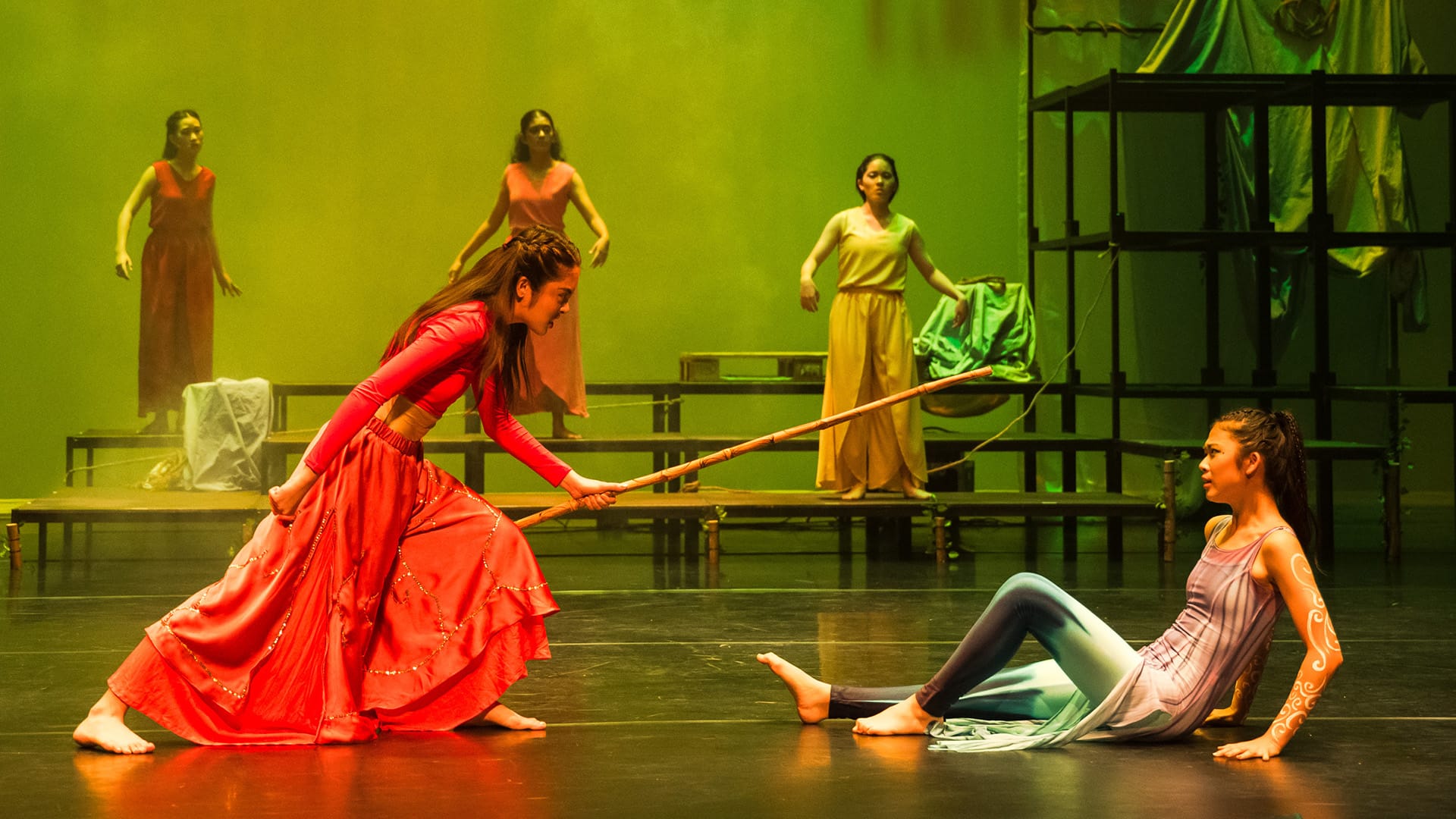 A performer in a red dress points a bamboo stick at a seated dancer on stage, with three others standing behind under green lighting.