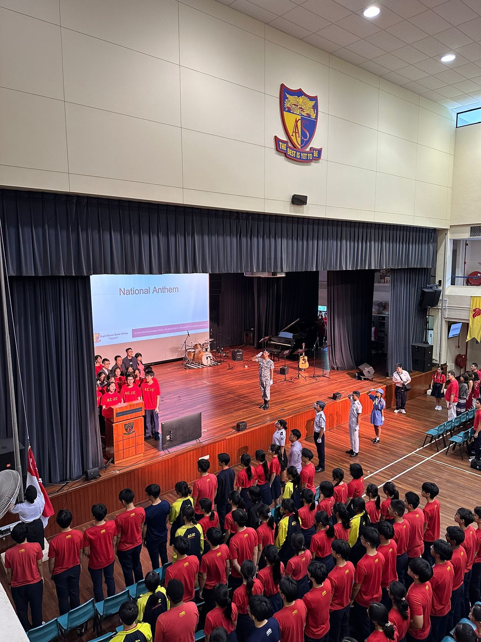 Students in red shirts stand in a school hall during a National Anthem ceremony, with a crest reading "The Best Is Yet To Be."
