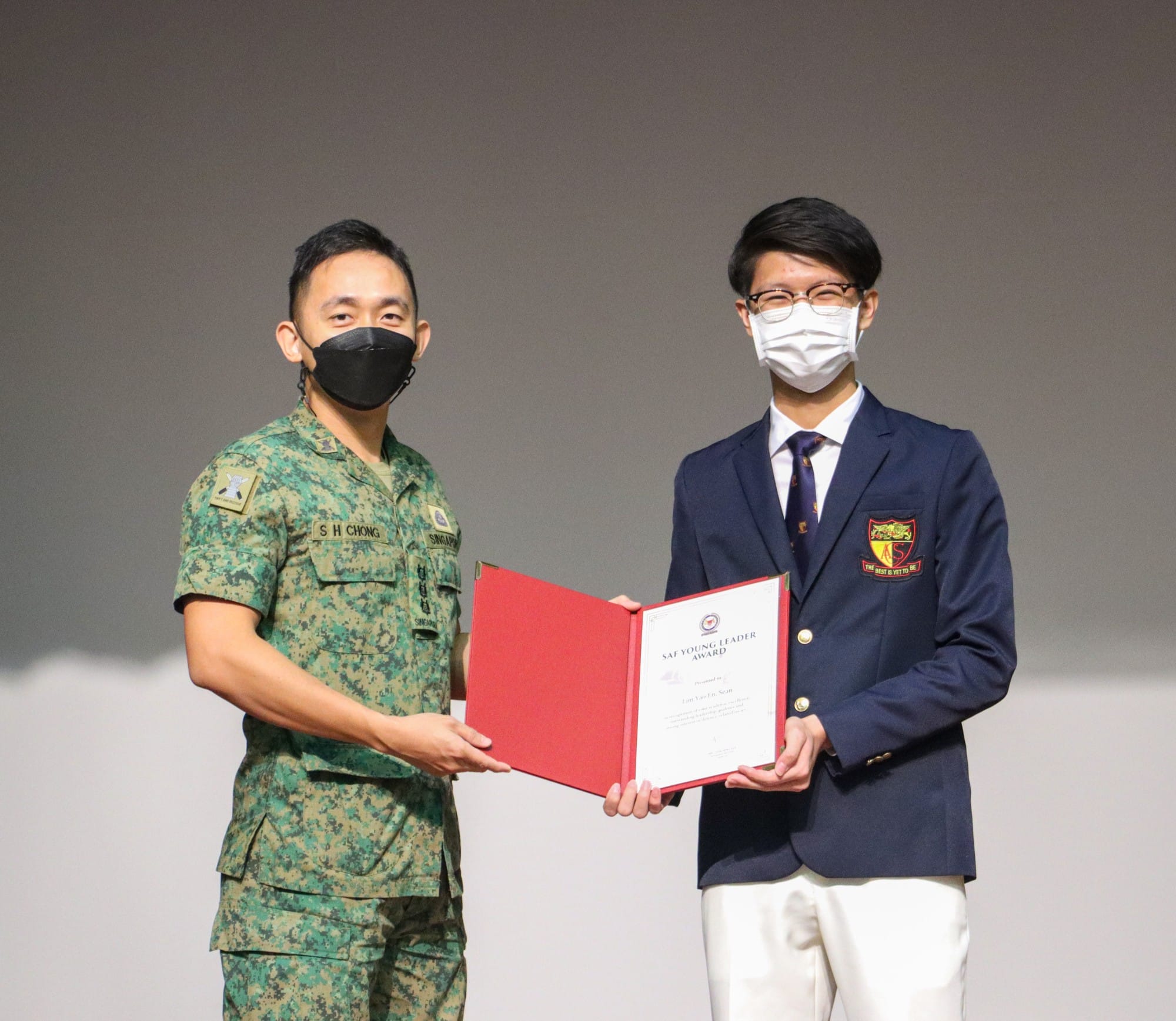 A soldier and a student in a blazer hold an SAF Young Leader Award certificate together, both wearing face masks.