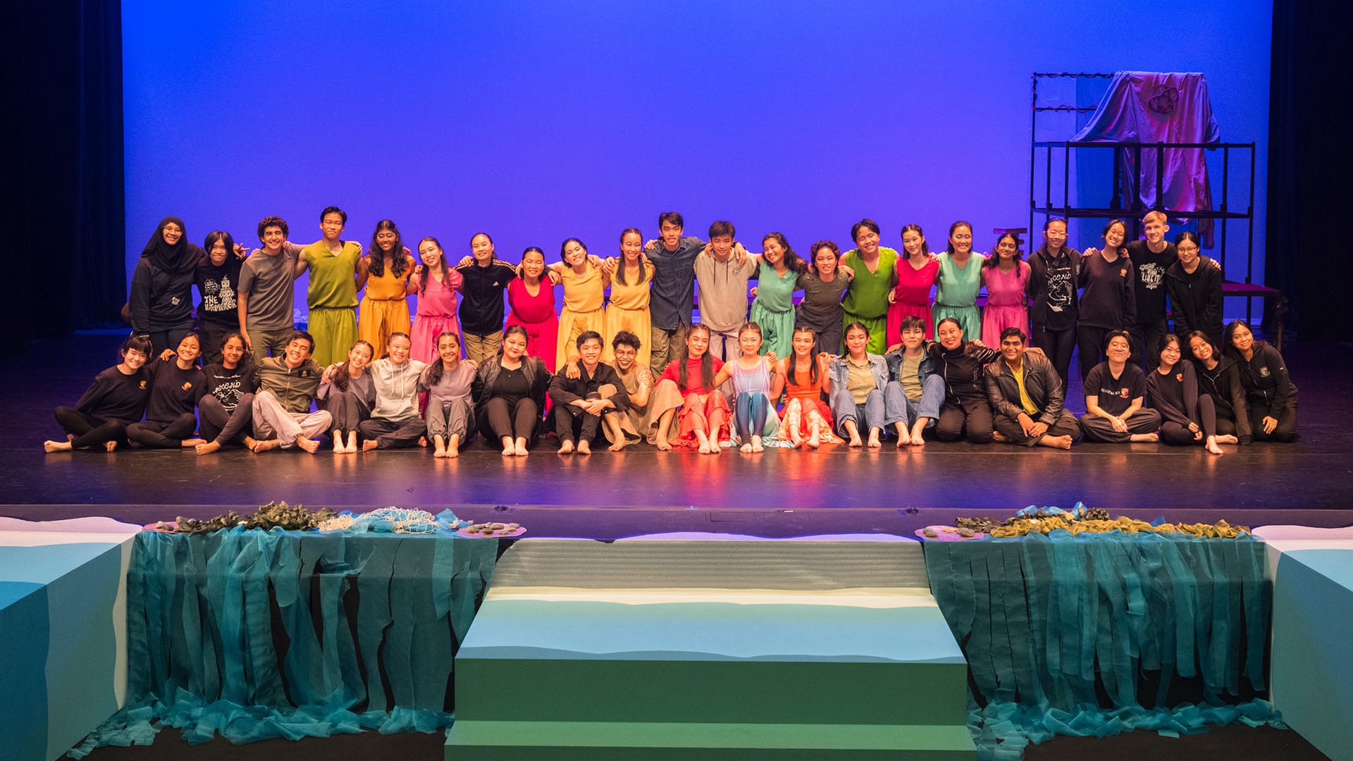 Large group of performers posing on a stage with blue backdrop, some in colorful costumes, others in black.