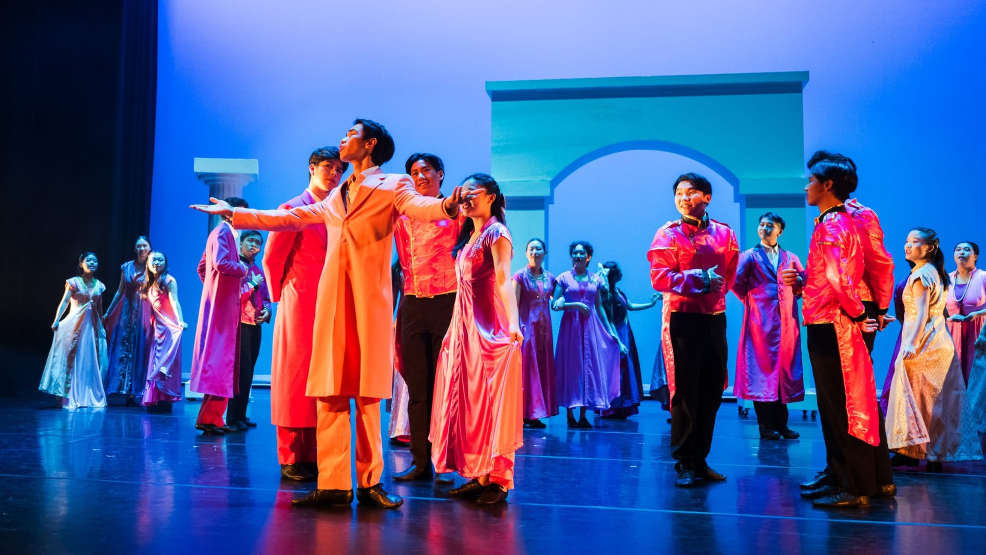 A large cast in colorful costumes performs on a blue-lit stage with an arched backdrop.