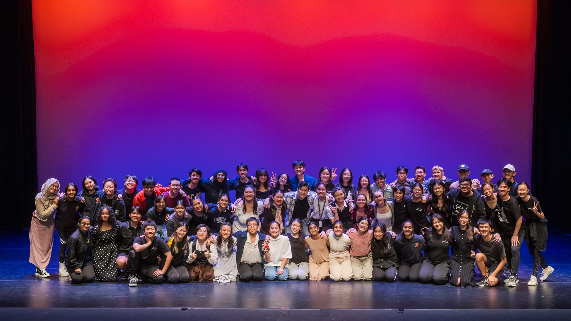Large group of young people posing together on a stage with a colorful red, pink, and purple lit backdrop.