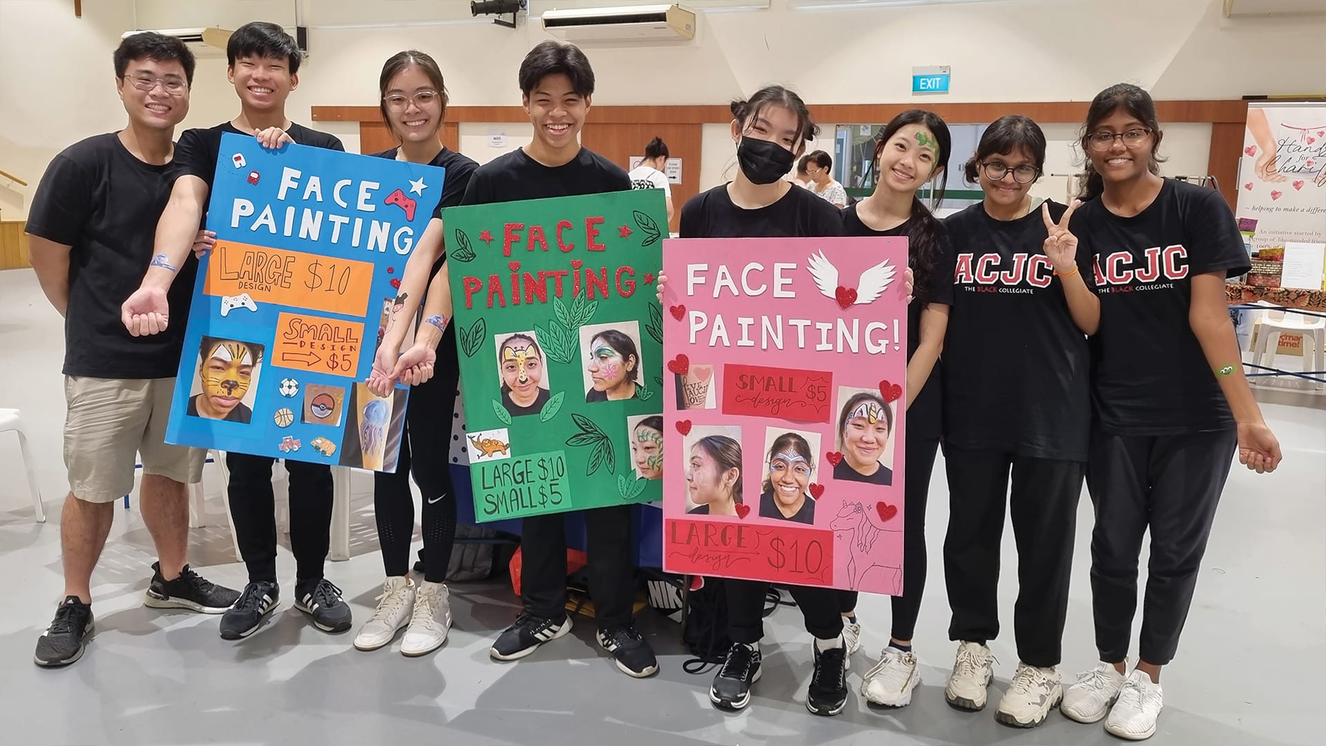 Eight smiling students in black shirts hold three colorful face painting signs showing prices of $5 and $10.