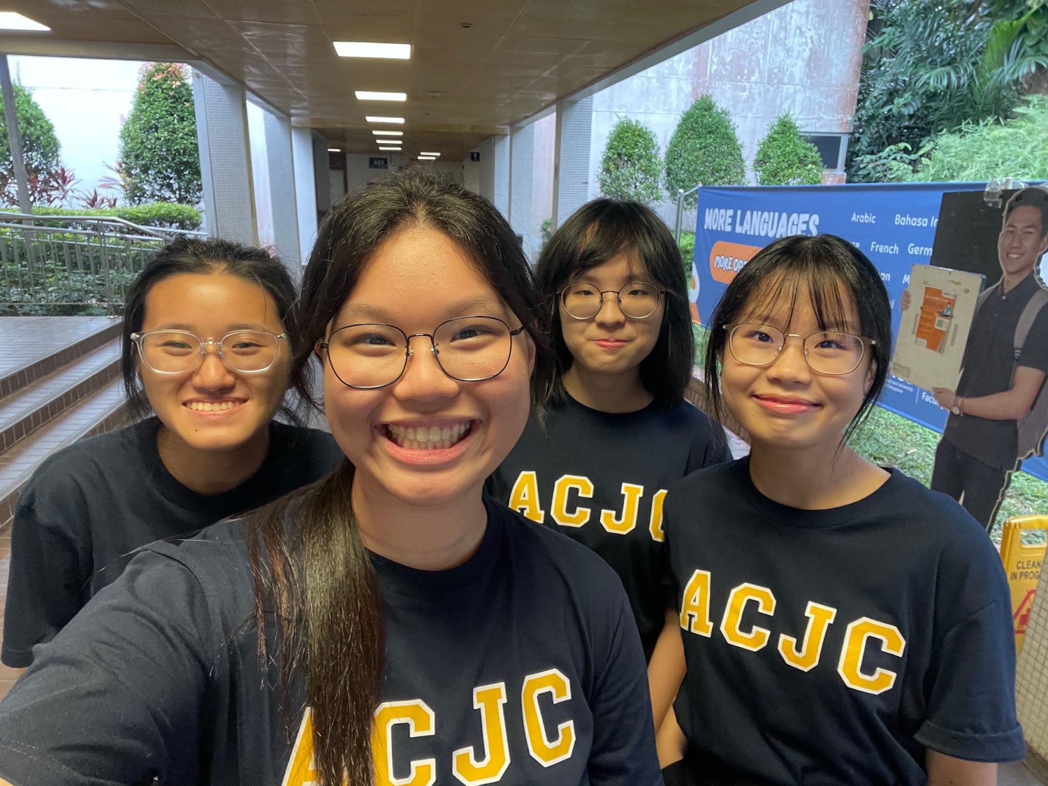 Four young women wearing black ACJC shirts smile for a selfie in an indoor corridor with greenery outside.