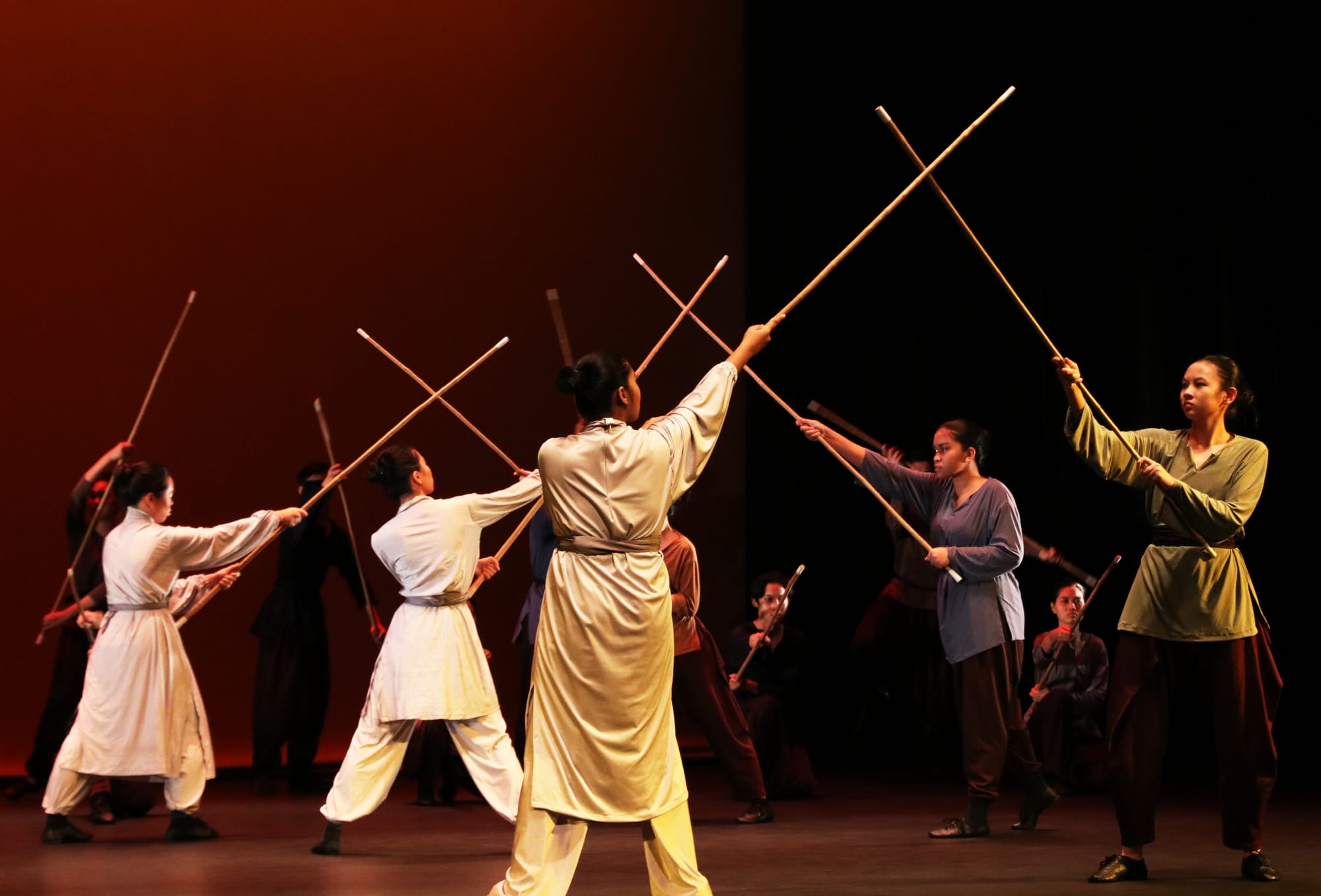 Performers in traditional robes cross long wooden staffs on a dimly lit stage during a martial arts dance performance.