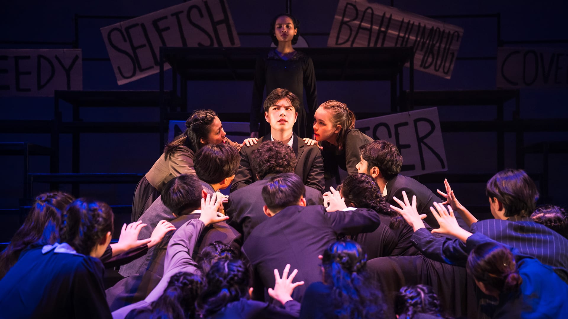A stage performance where a crowd surrounds a young man as signs reading "Selfish" and "Bah Humbug" hang behind them.