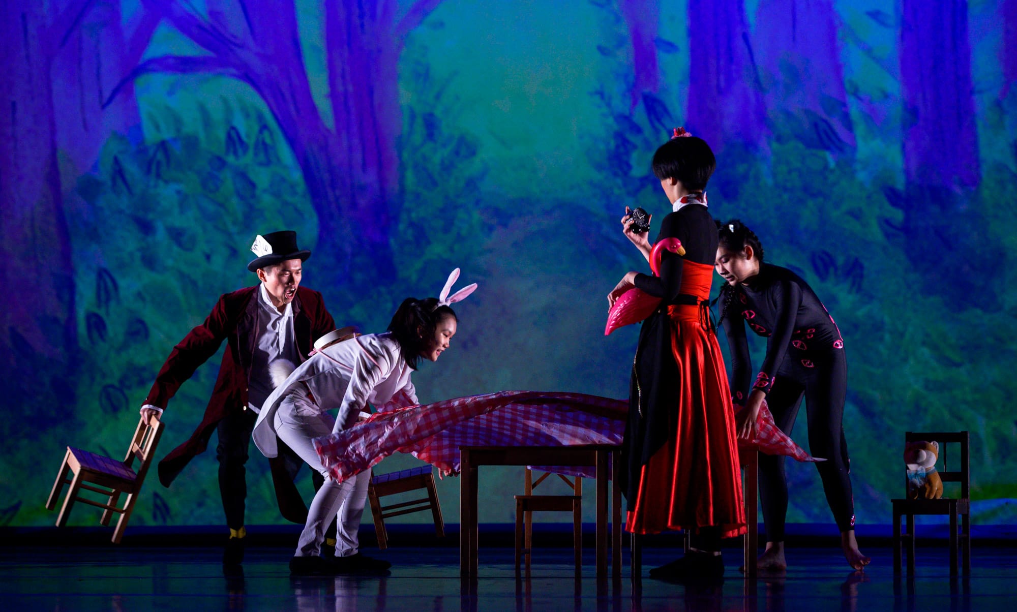Four performers in Alice in Wonderland costumes act on a stage with a colorful forest backdrop and a table.