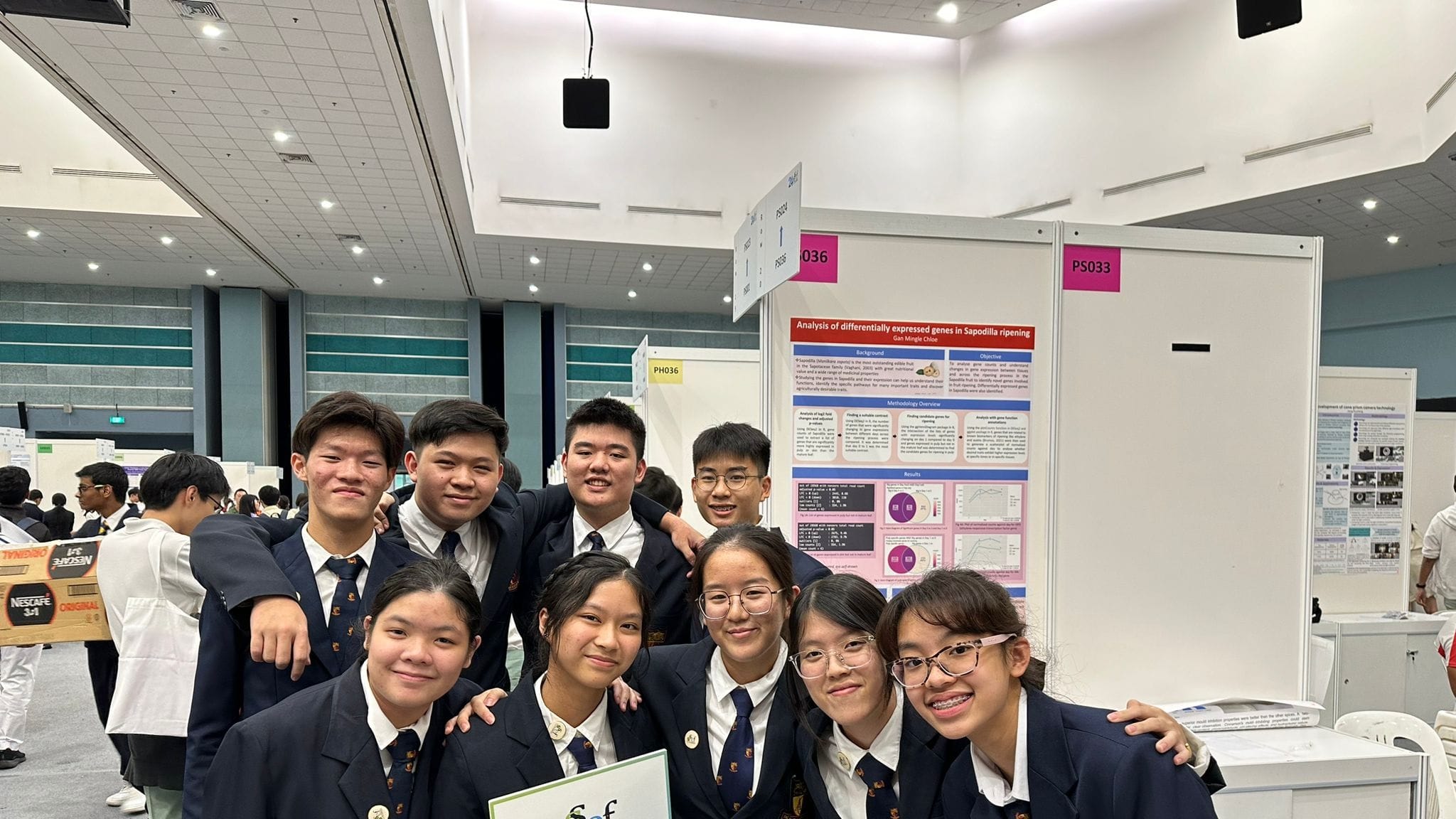 Nine students in dark blazers pose together at a science fair, with research posters visible behind them.