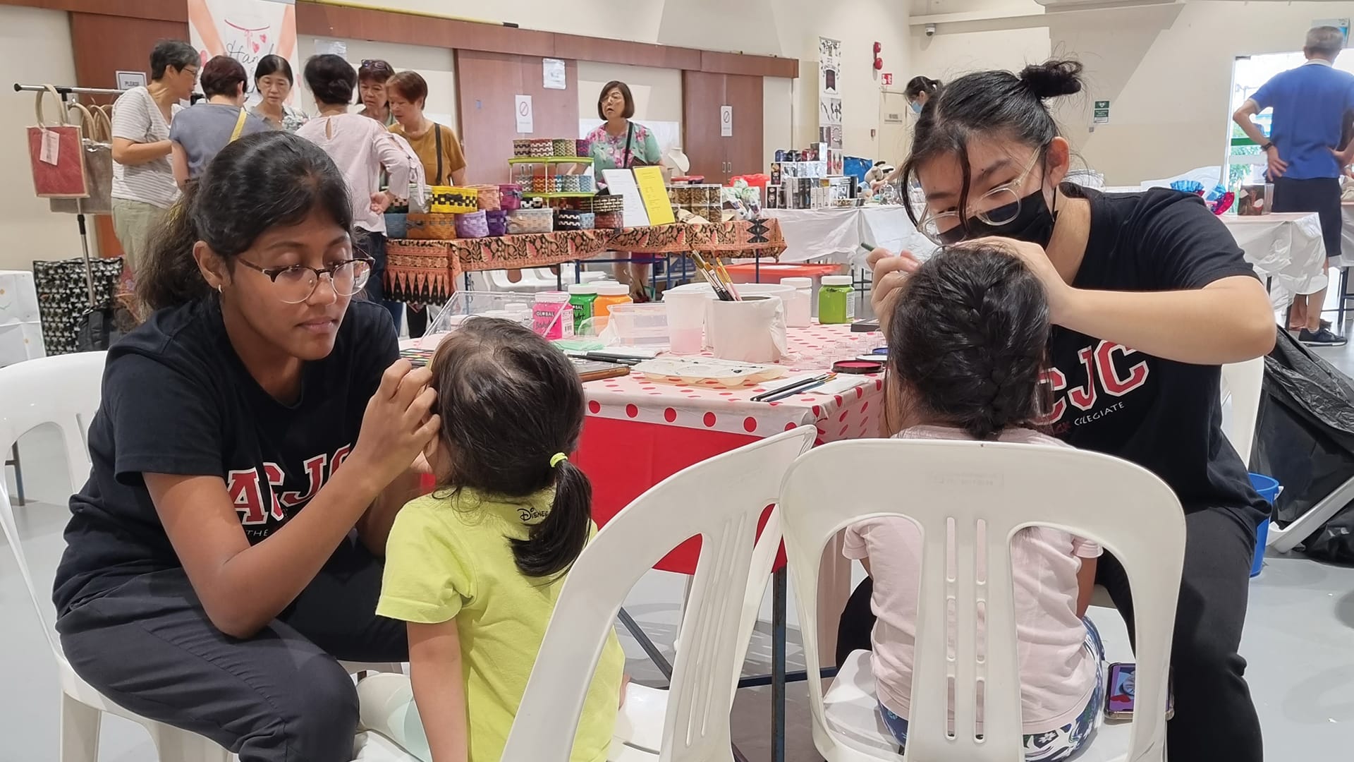 Two volunteers in black CJC shirts paint faces of young children at a community event with vendor stalls behind.