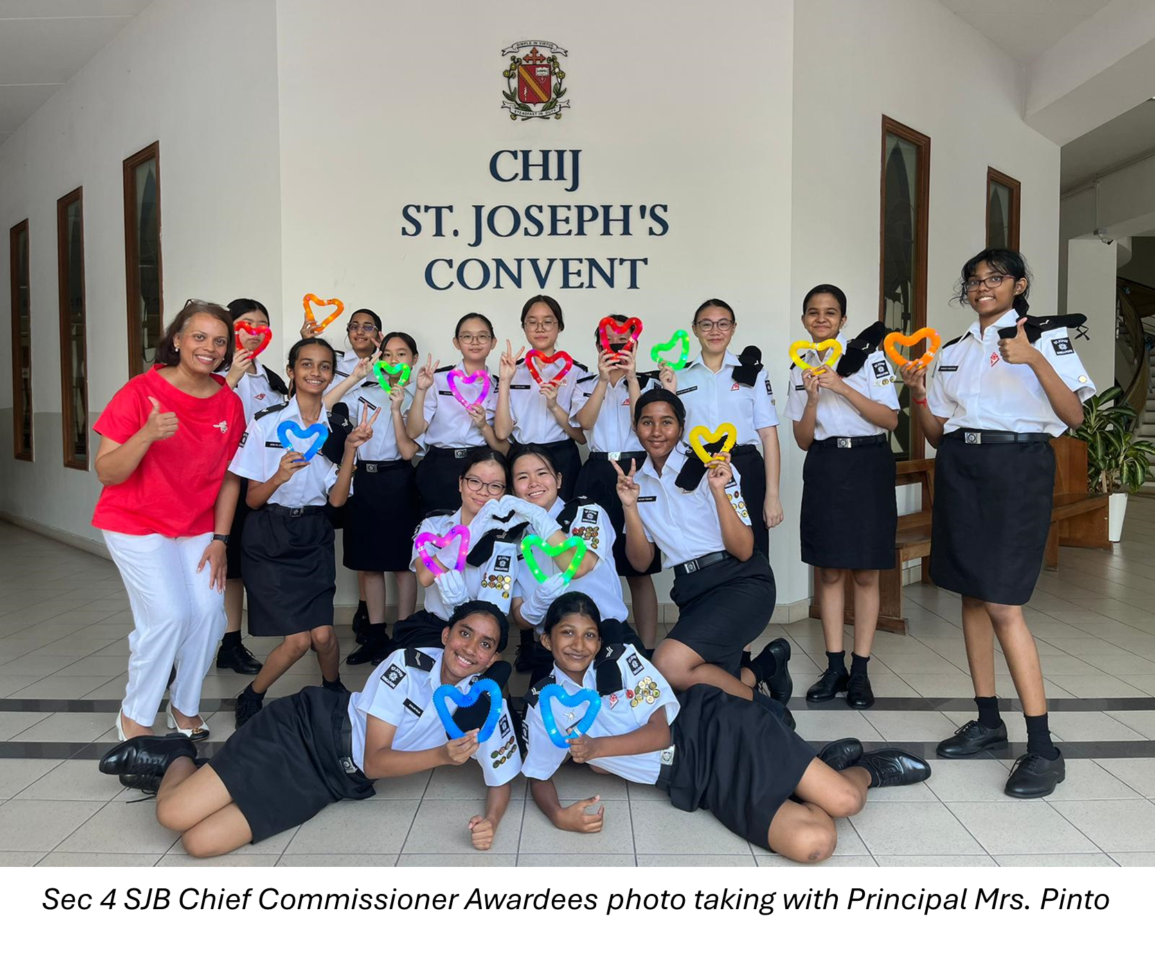 Group of uniformed school girls holding colorful heart shapes pose with a woman in red at CHIJ St. Joseph's Convent.