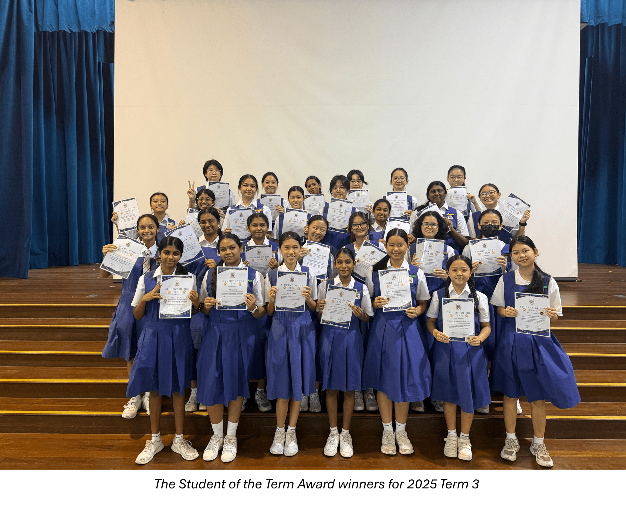 Group of schoolgirls in blue uniforms holding Student of the Term Award certificates on a school stage.