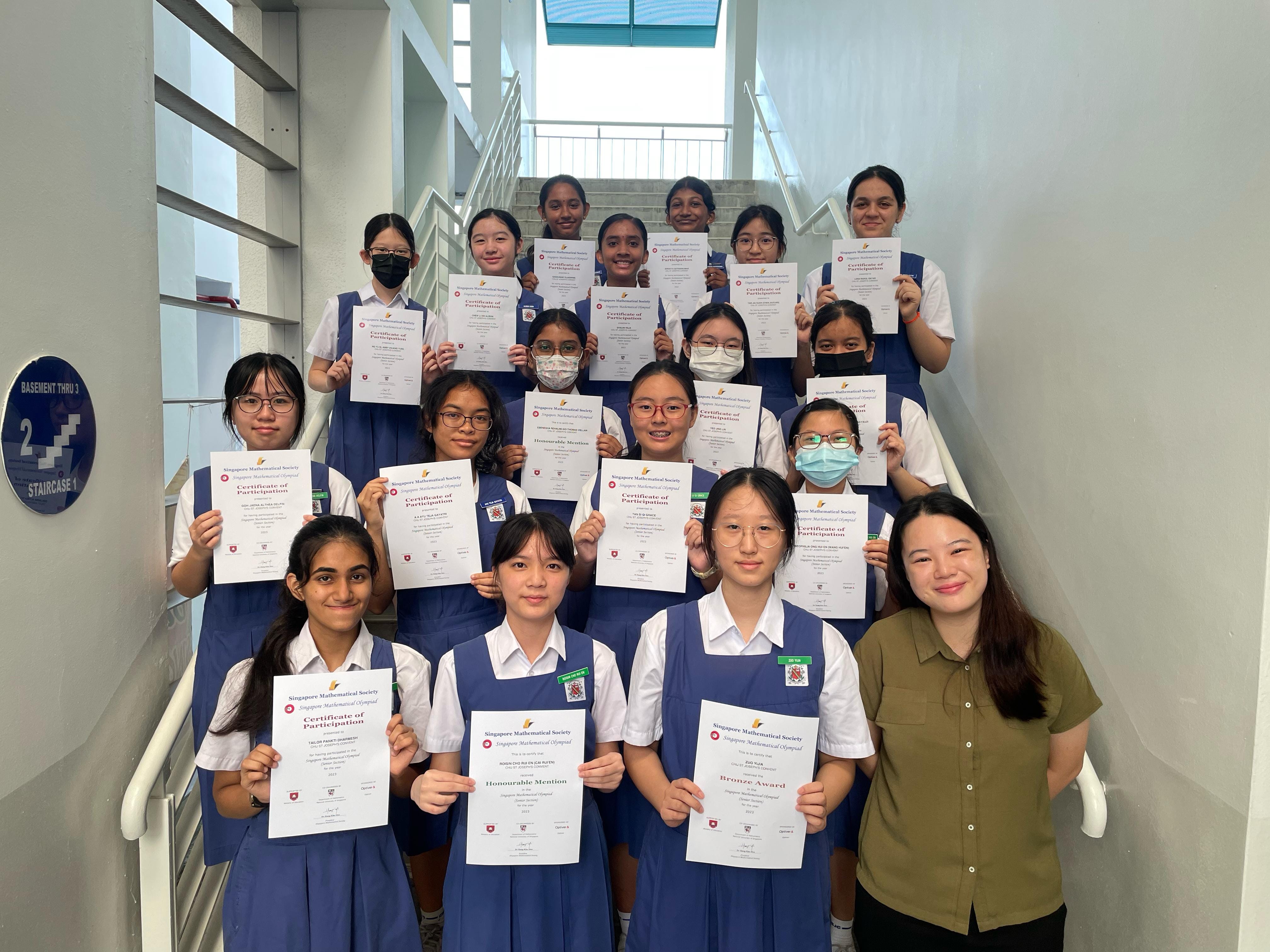 A group of school girls in blue uniforms hold Singapore Mathematical Olympiad certificates on a staircase.