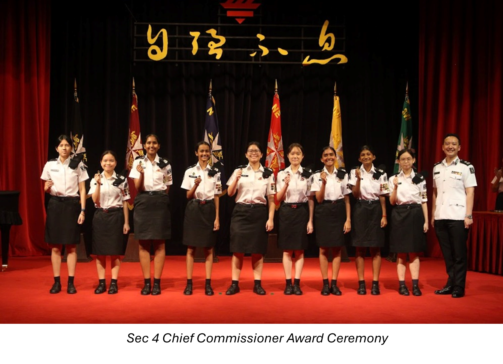 Ten uniformed students and one adult officer stand on a red stage holding awards, with flags behind them.