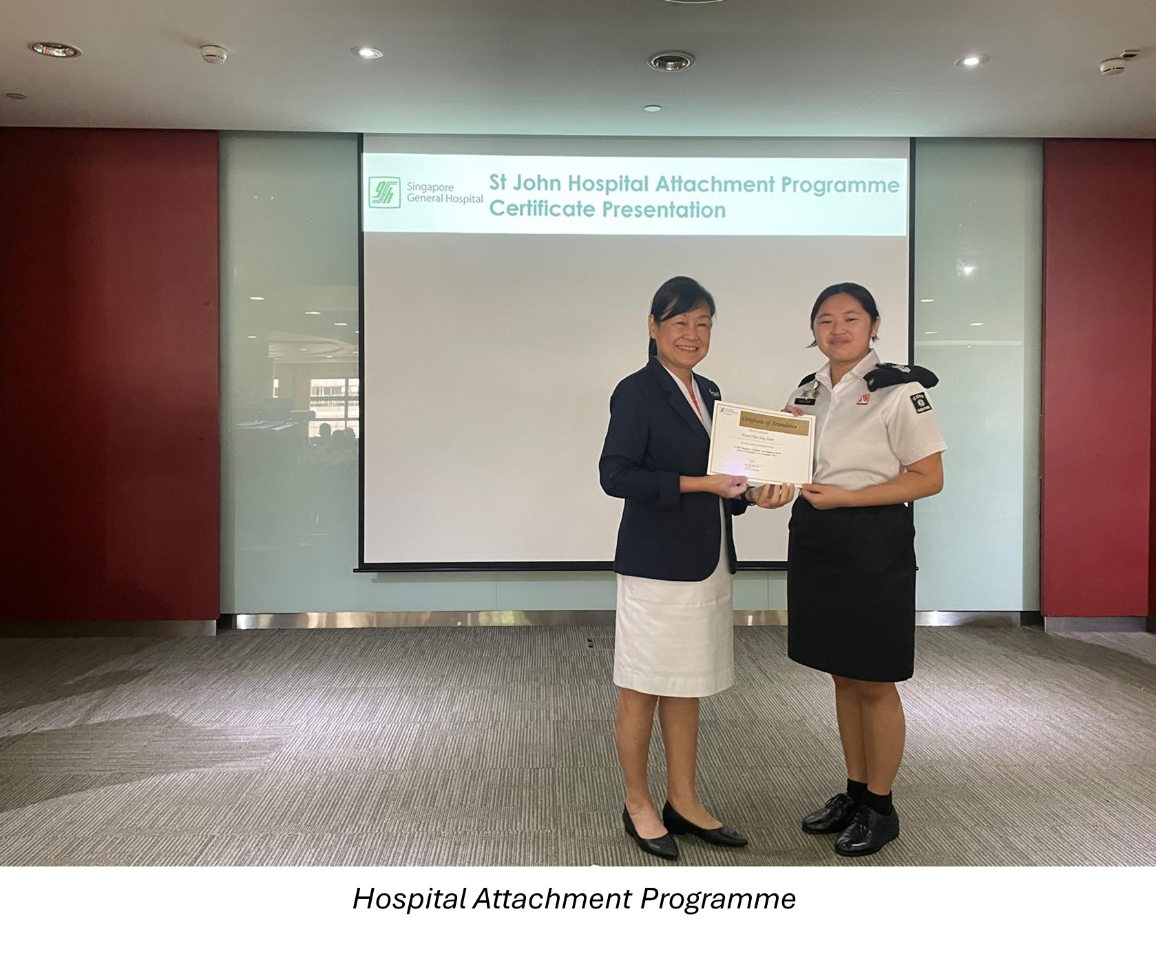 Two women hold a certificate in front of a screen showing St John Hospital Attachment Programme Certificate Presentation.