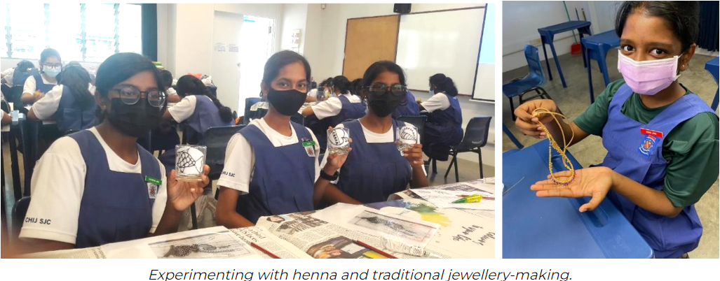 Students in blue aprons and masks hold henna-decorated jars and beaded jewellery during a craft activity in a classroom.