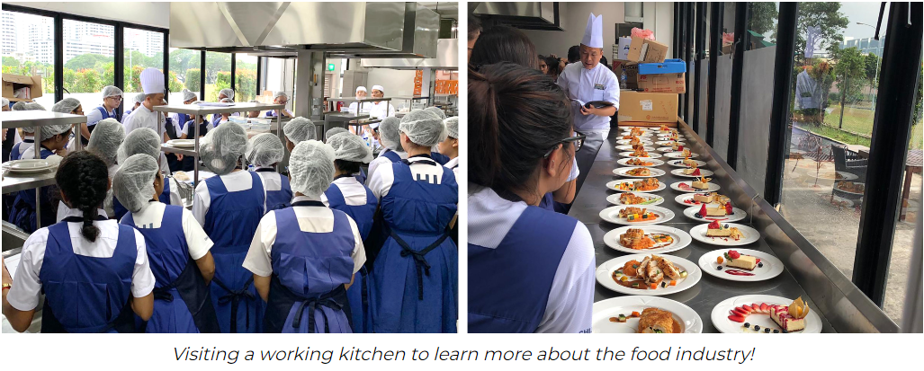 Students in blue aprons and hairnets observe a chef in a professional kitchen with plated dishes lined up on a counter.