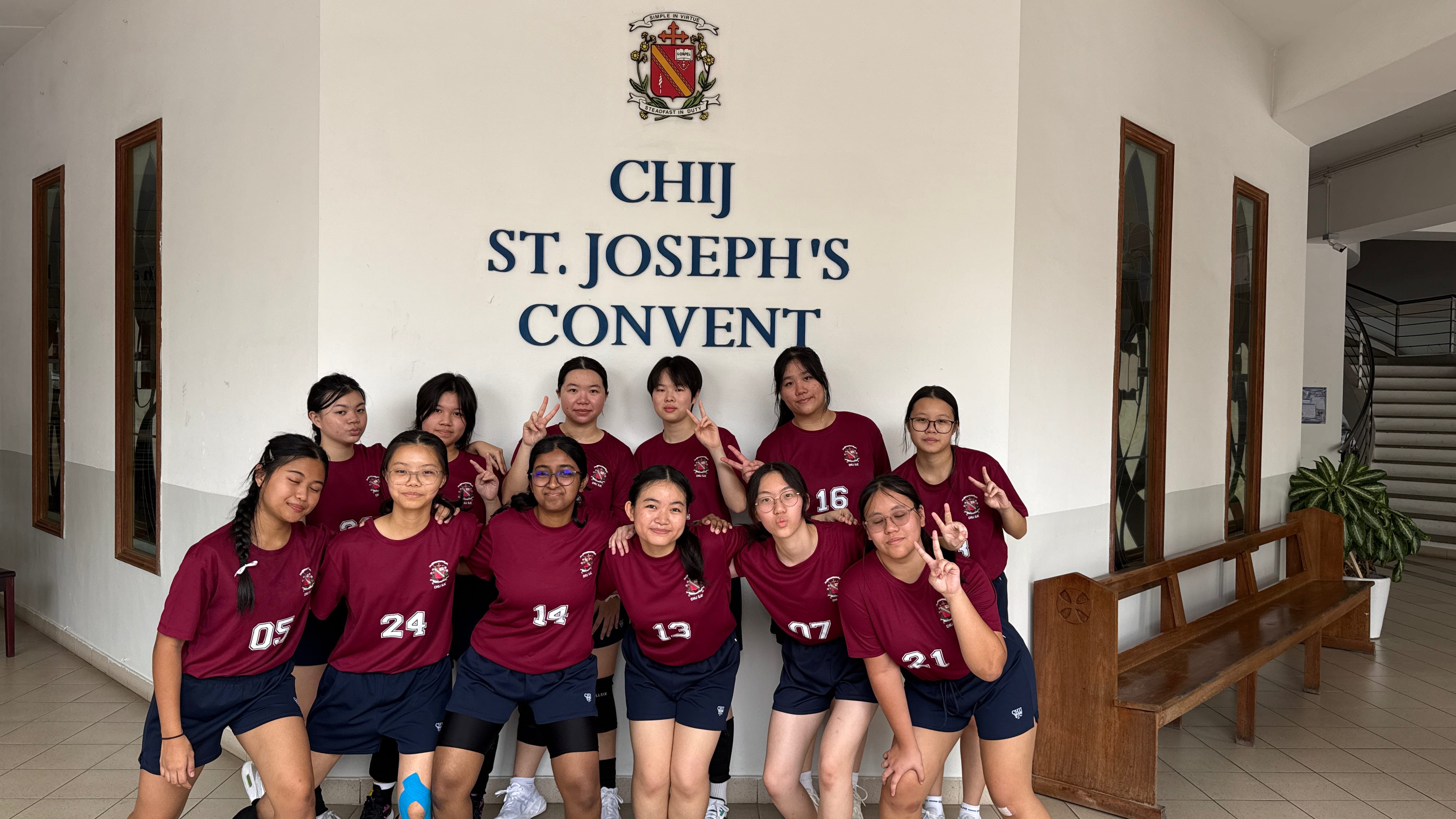 A group of girls in maroon numbered jerseys pose with peace signs in front of a CHIJ St. Joseph's Convent sign.