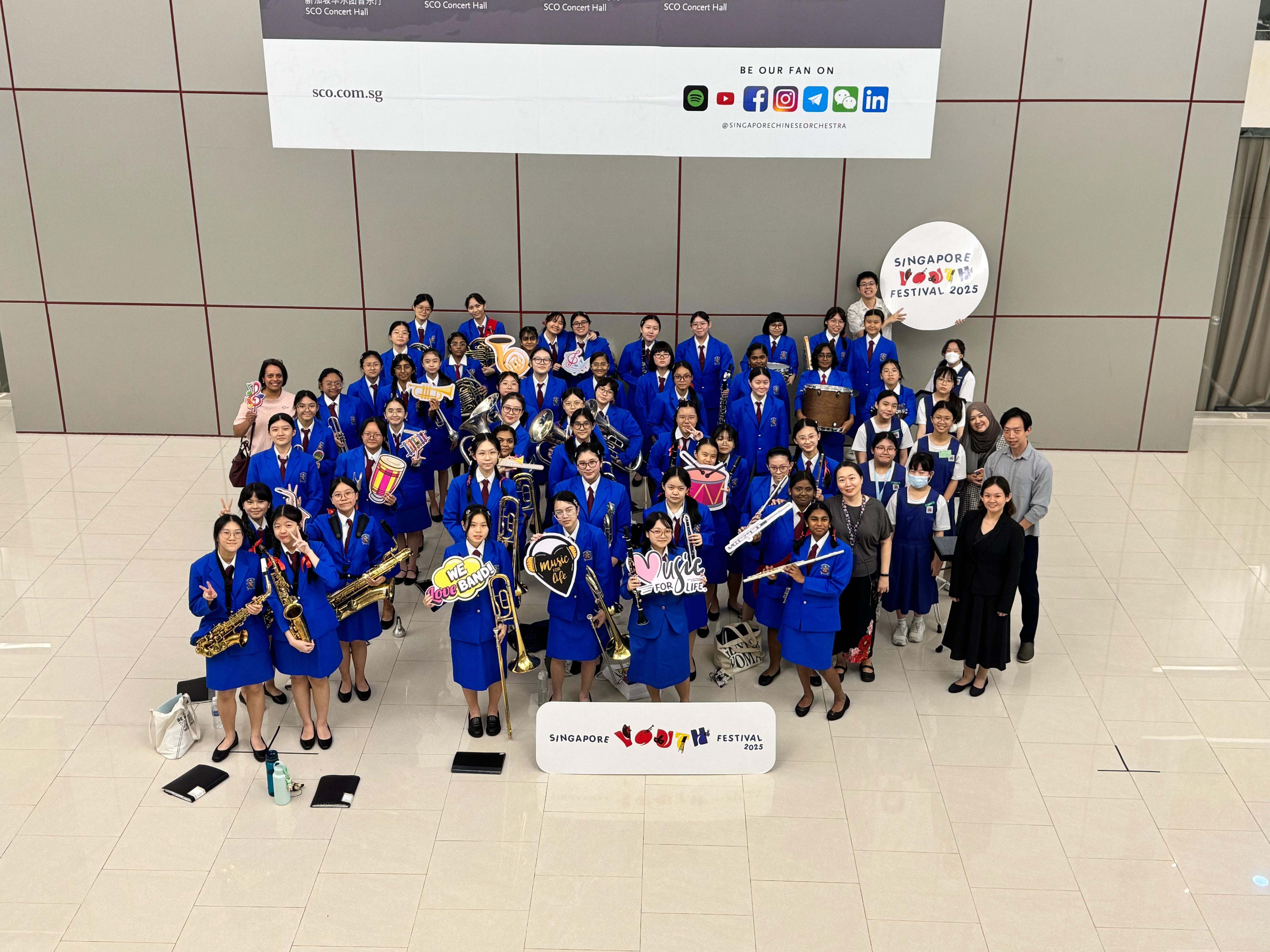 Large group of students in blue uniforms holding instruments and signs at Singapore Youth Festival 2025 inside SCO Concert Hall.