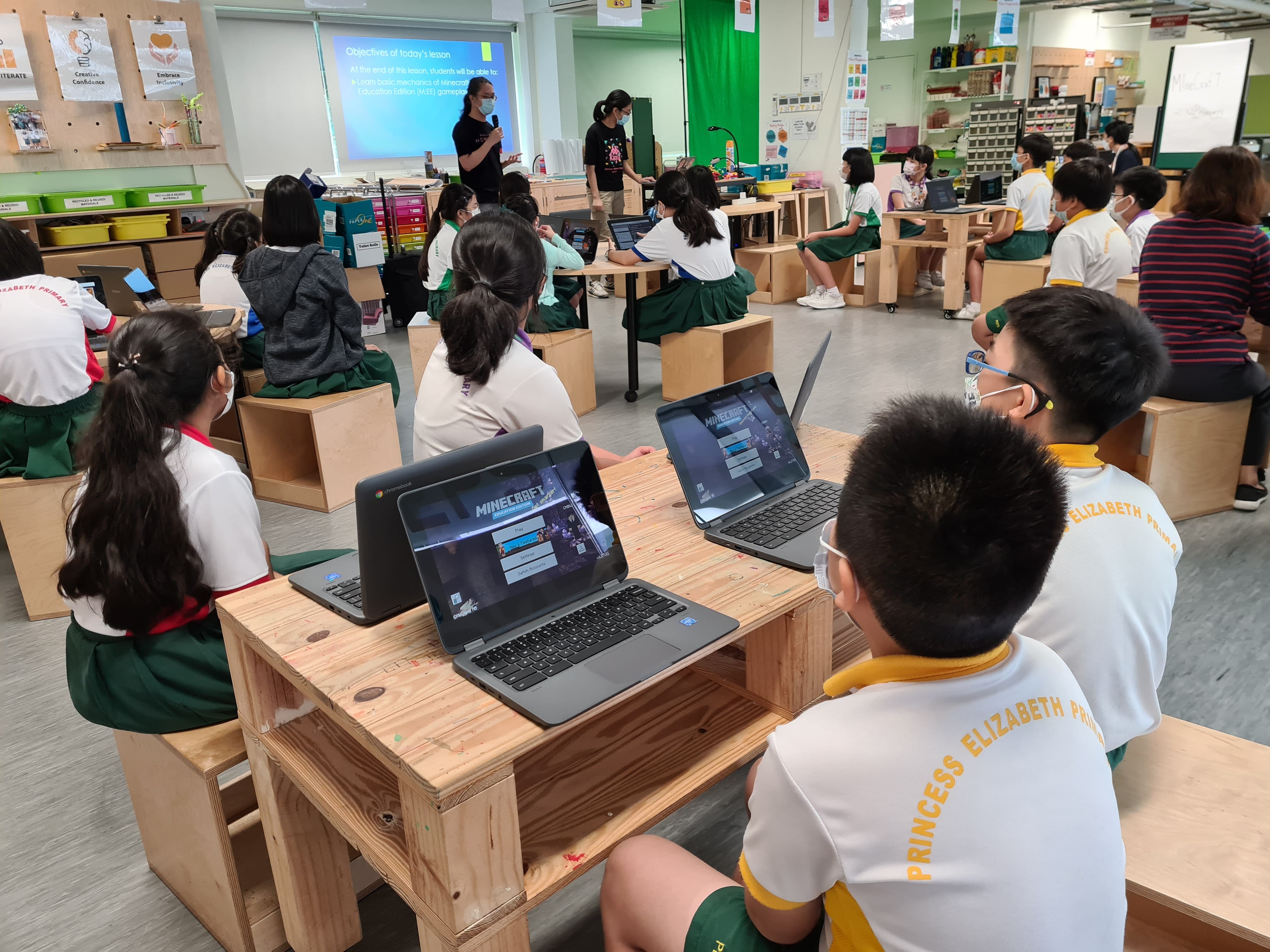 Students in uniforms sit at wooden desks with Minecraft open on laptops, facing a teacher presenting slides.