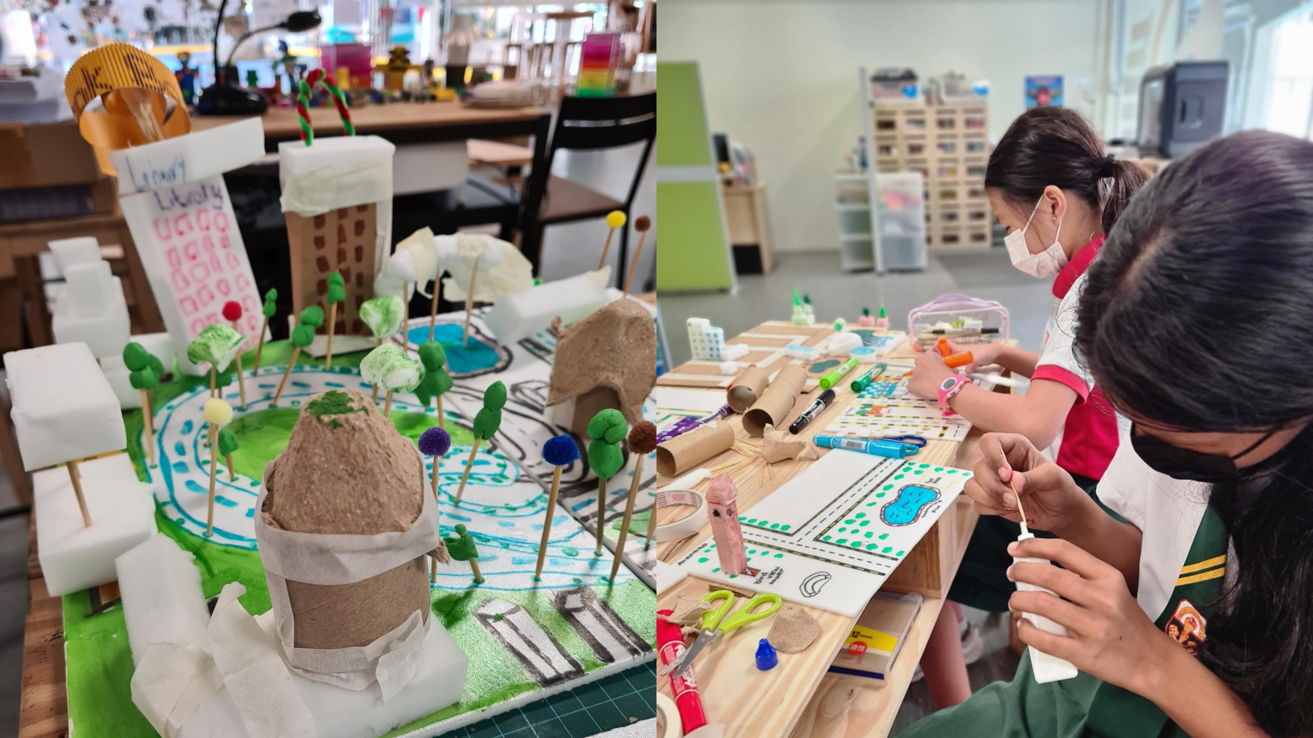 Two masked students build a colorful 3D town model using foam, cardboard tubes, pom-poms, and markers.
