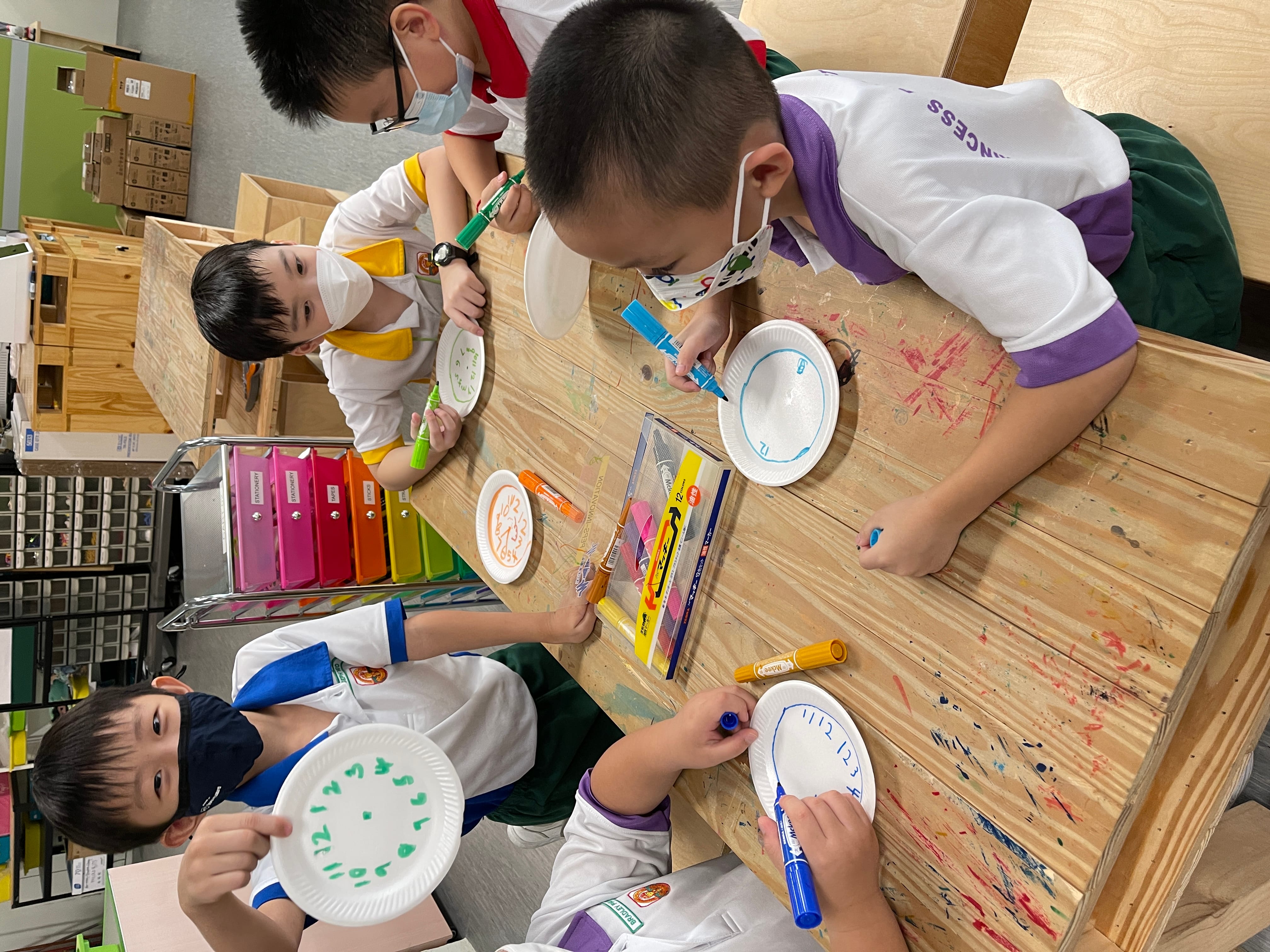 Four masked children at a paint-splattered table drawing with markers on paper plates in a classroom.