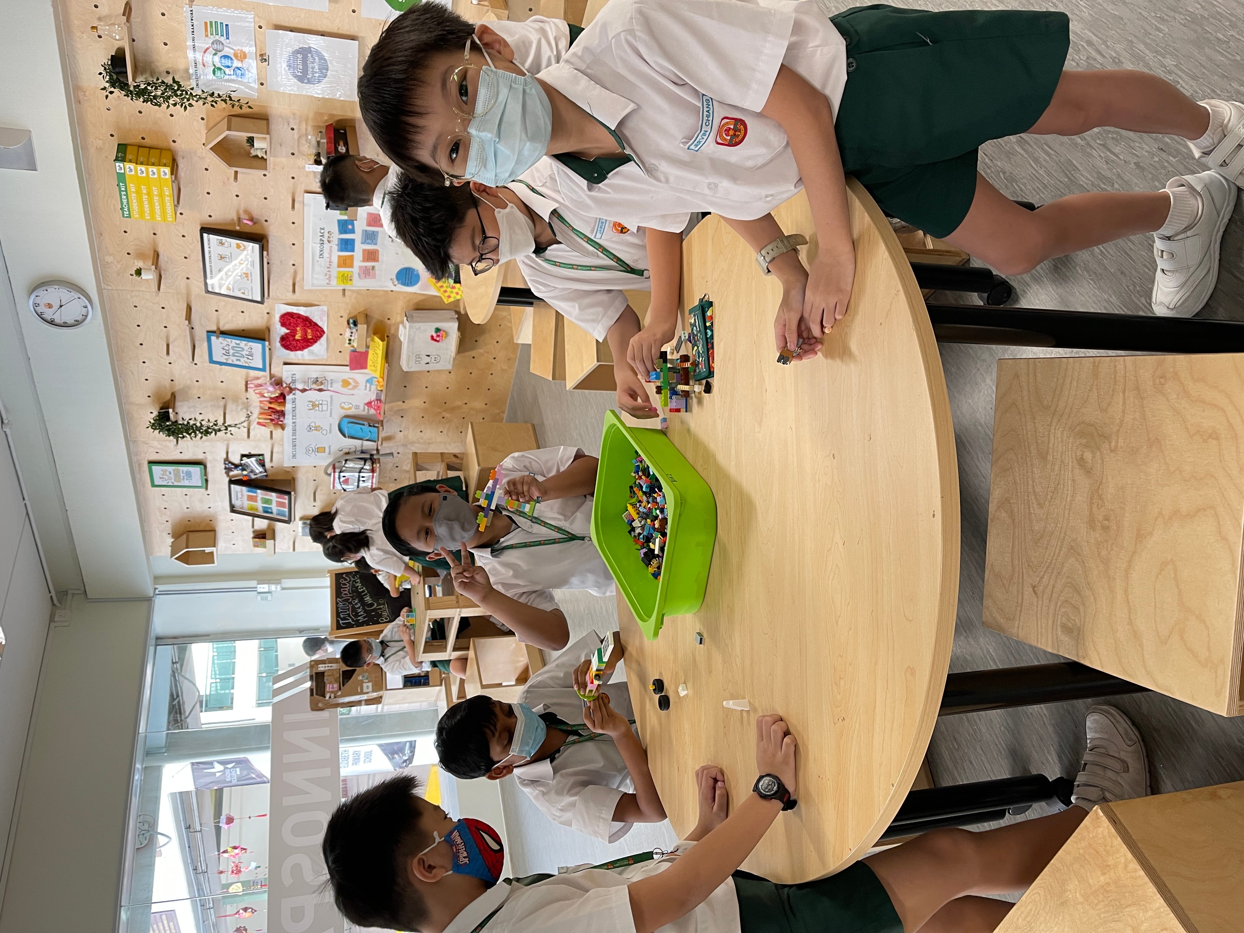 Young students in uniforms and face masks build with colorful bricks around a round wooden table in a bright classroom.