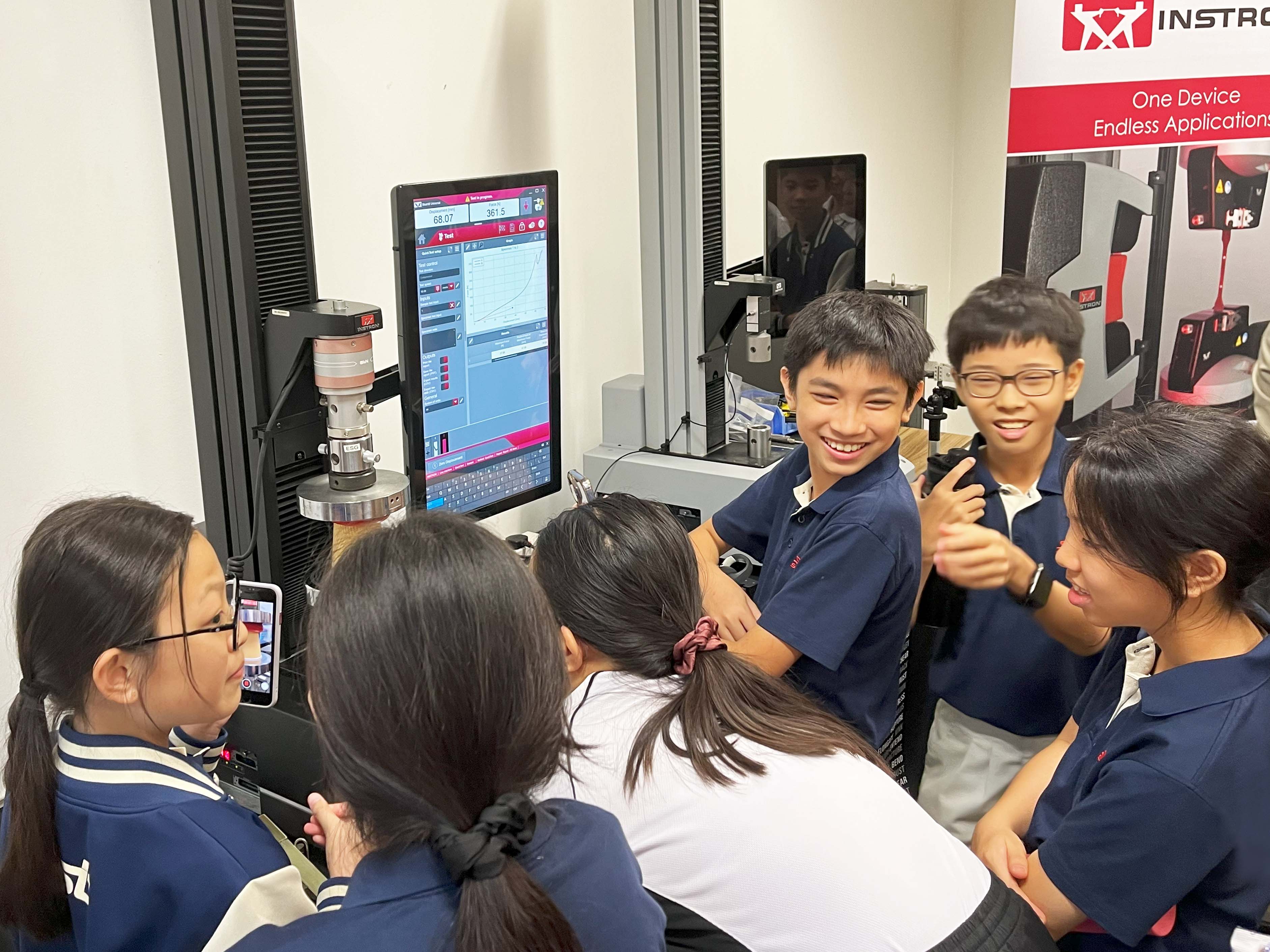 Several smiling students in navy uniforms gather around an Instron testing machine with a monitor showing test data.