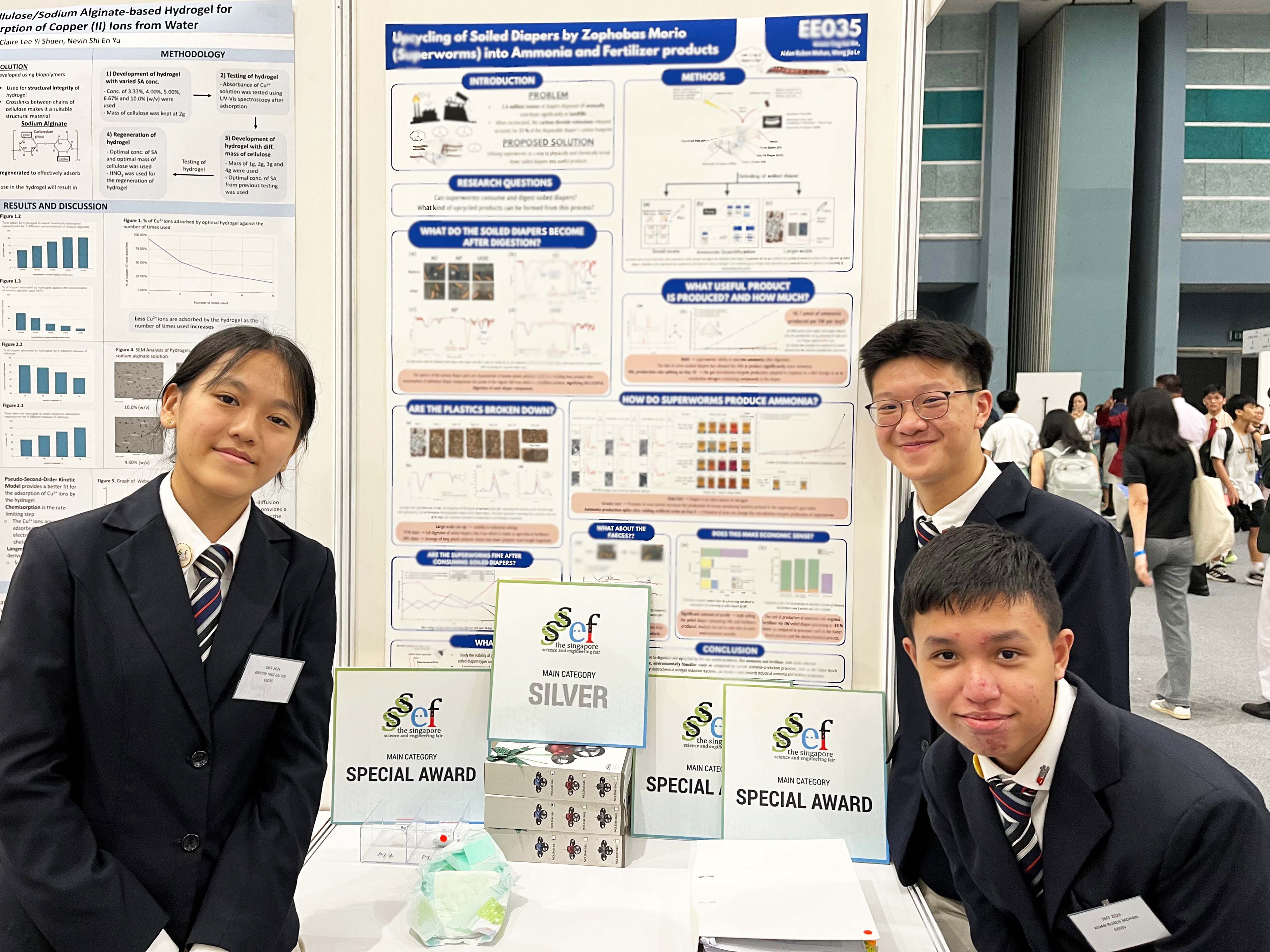 Three students in school uniforms pose at a science fair booth displaying Special Award and Silver award plaques.