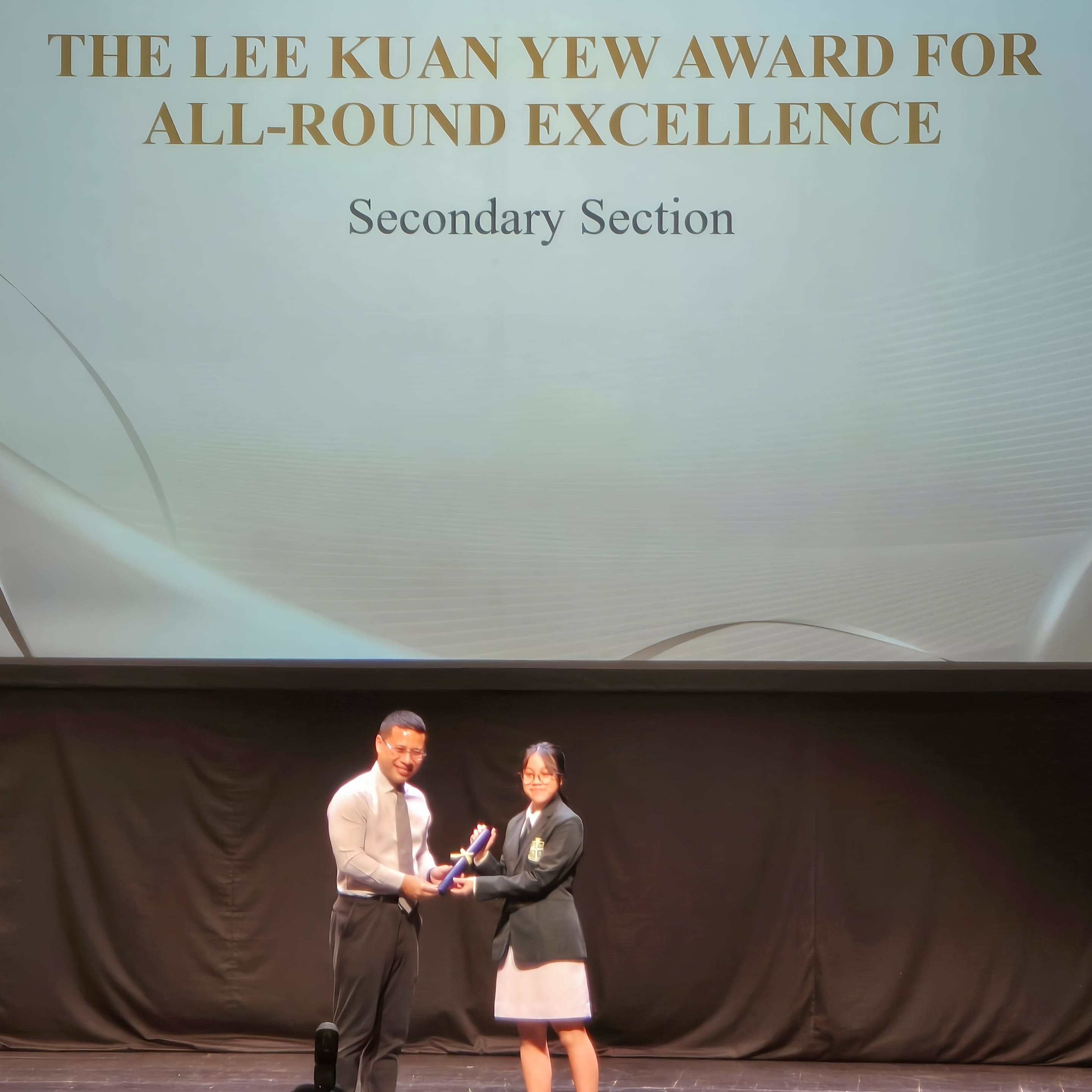 A man presents an award to a uniformed student on stage at the Lee Kuan Yew Award for All-Round Excellence ceremony.