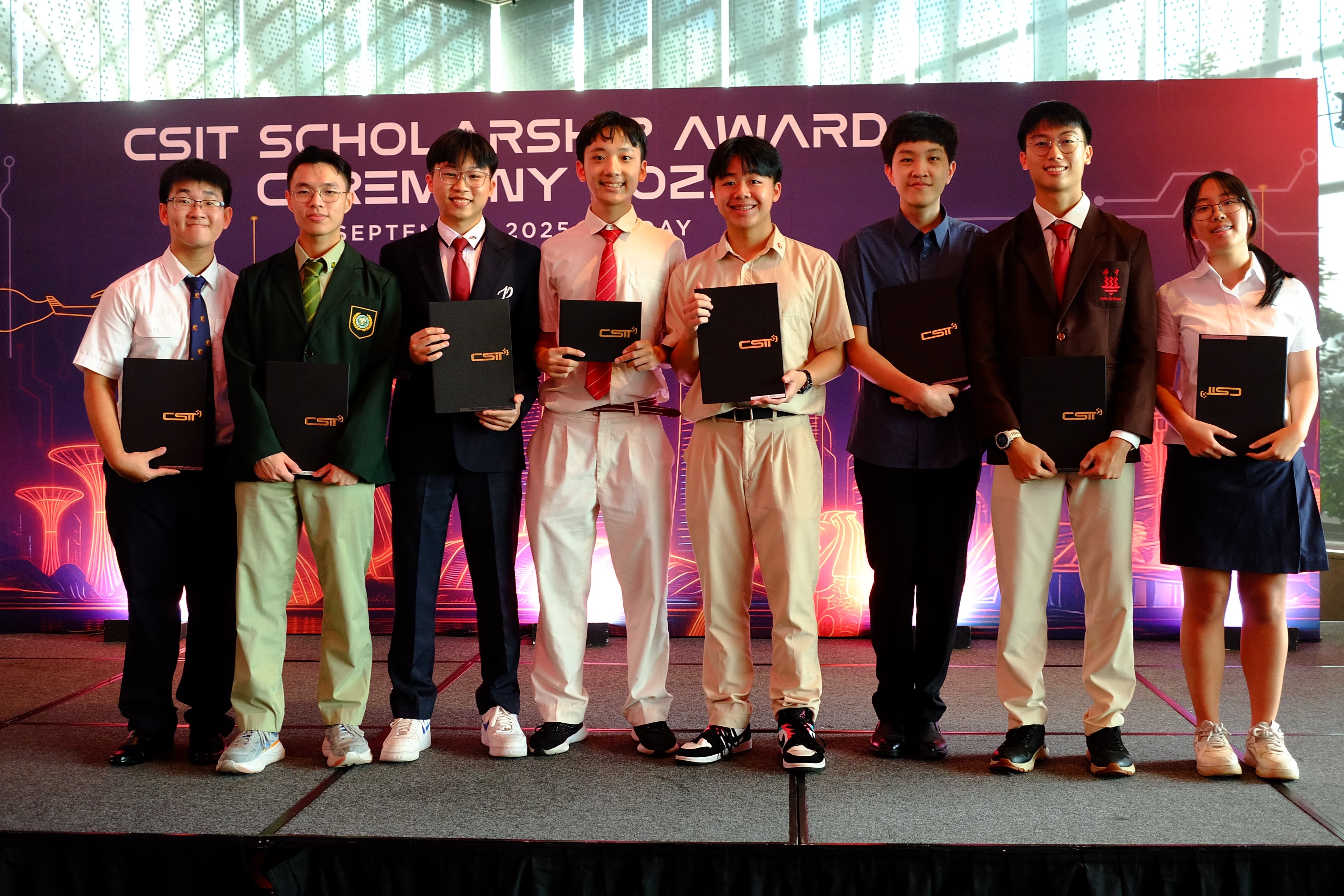 Eight young students stand on a stage holding black folders at the CSIT Scholarship Award Ceremony 2025.