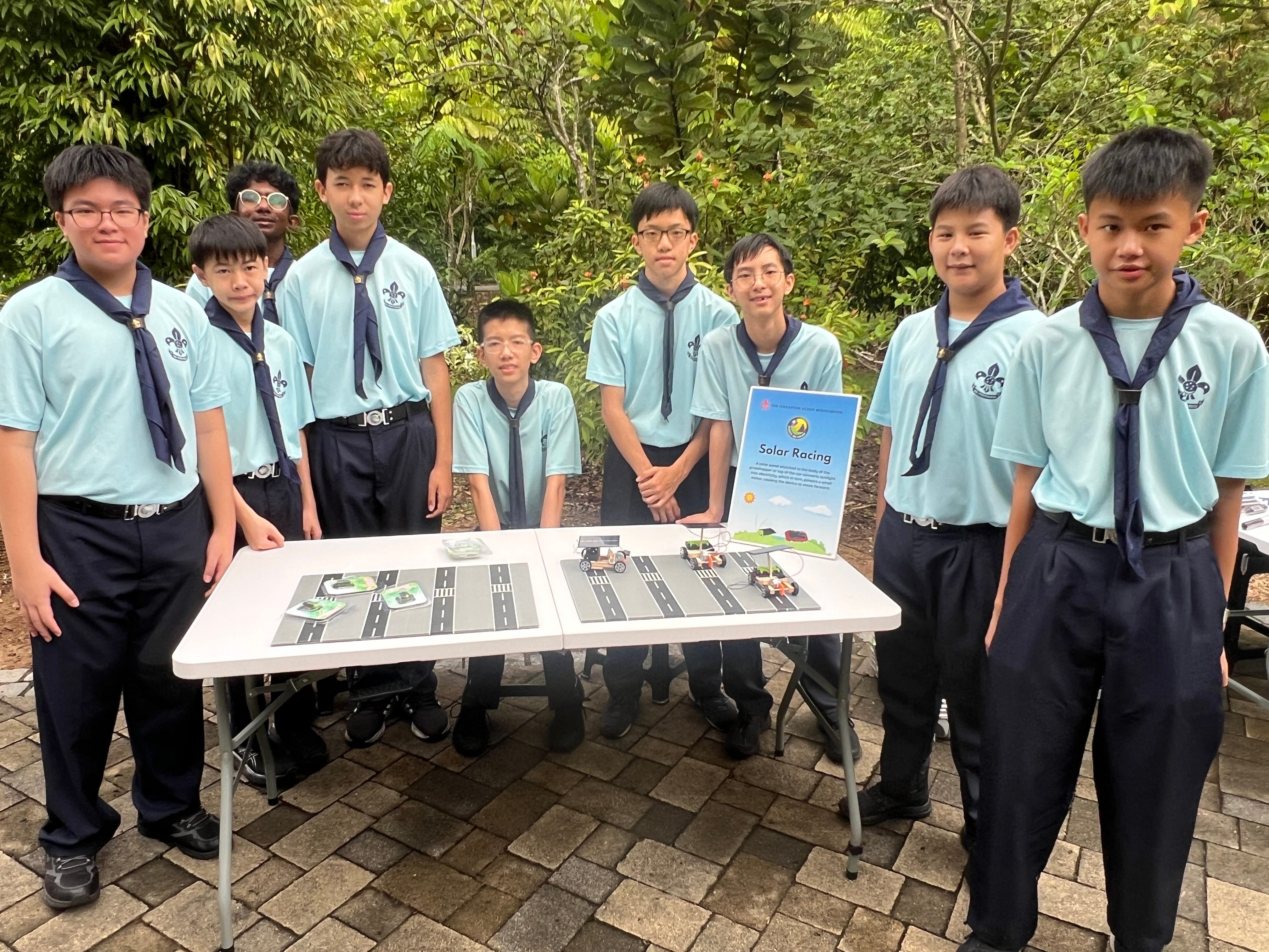 Nine scouts in light blue uniforms stand outdoors around a table displaying a Solar Racing project with small model cars.