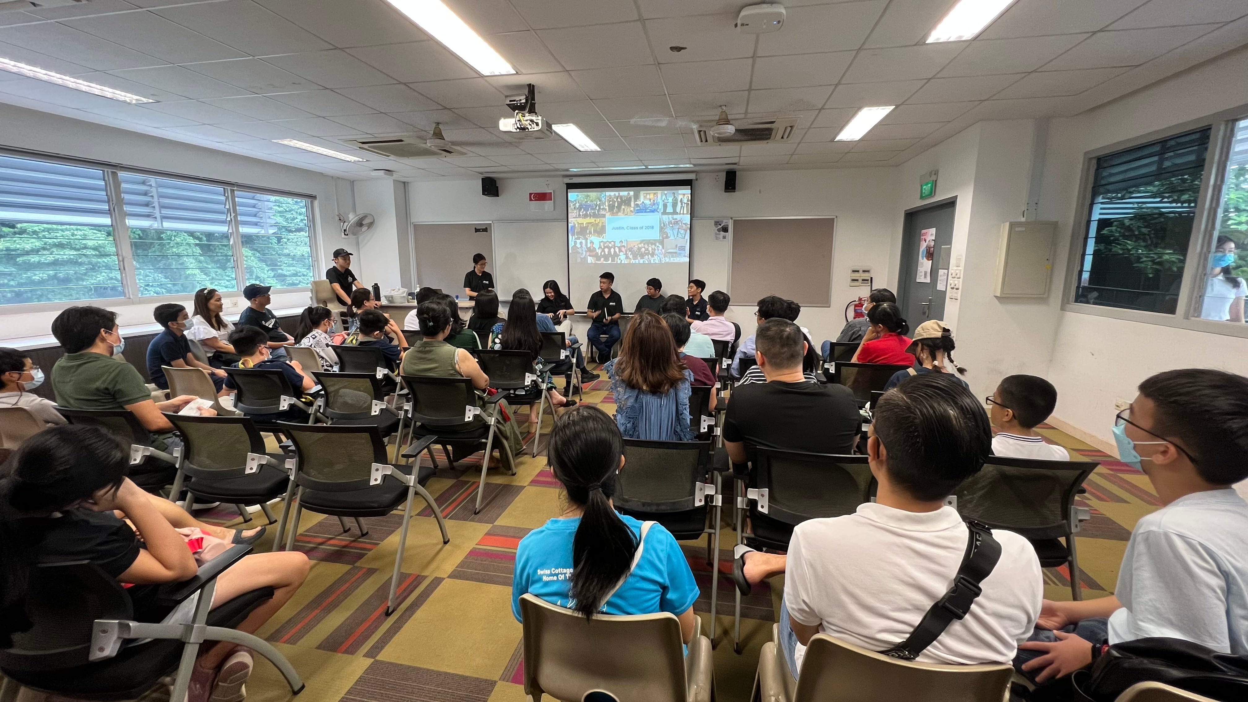 A group of people seated in a classroom watching a panel discussion with a projected slideshow at the front.