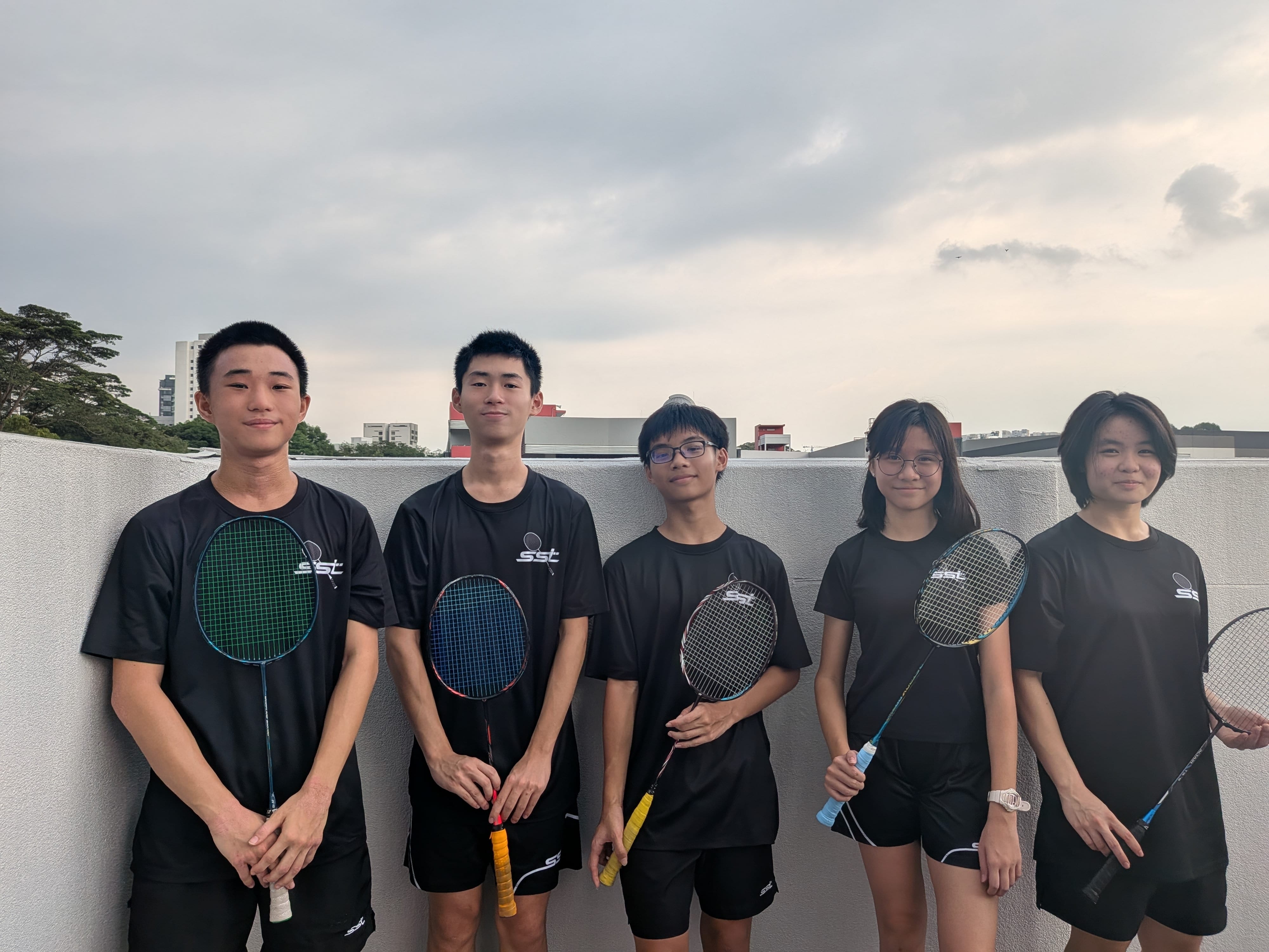 Five youths in black shirts pose with badminton rackets in front of a low wall under cloudy sky.