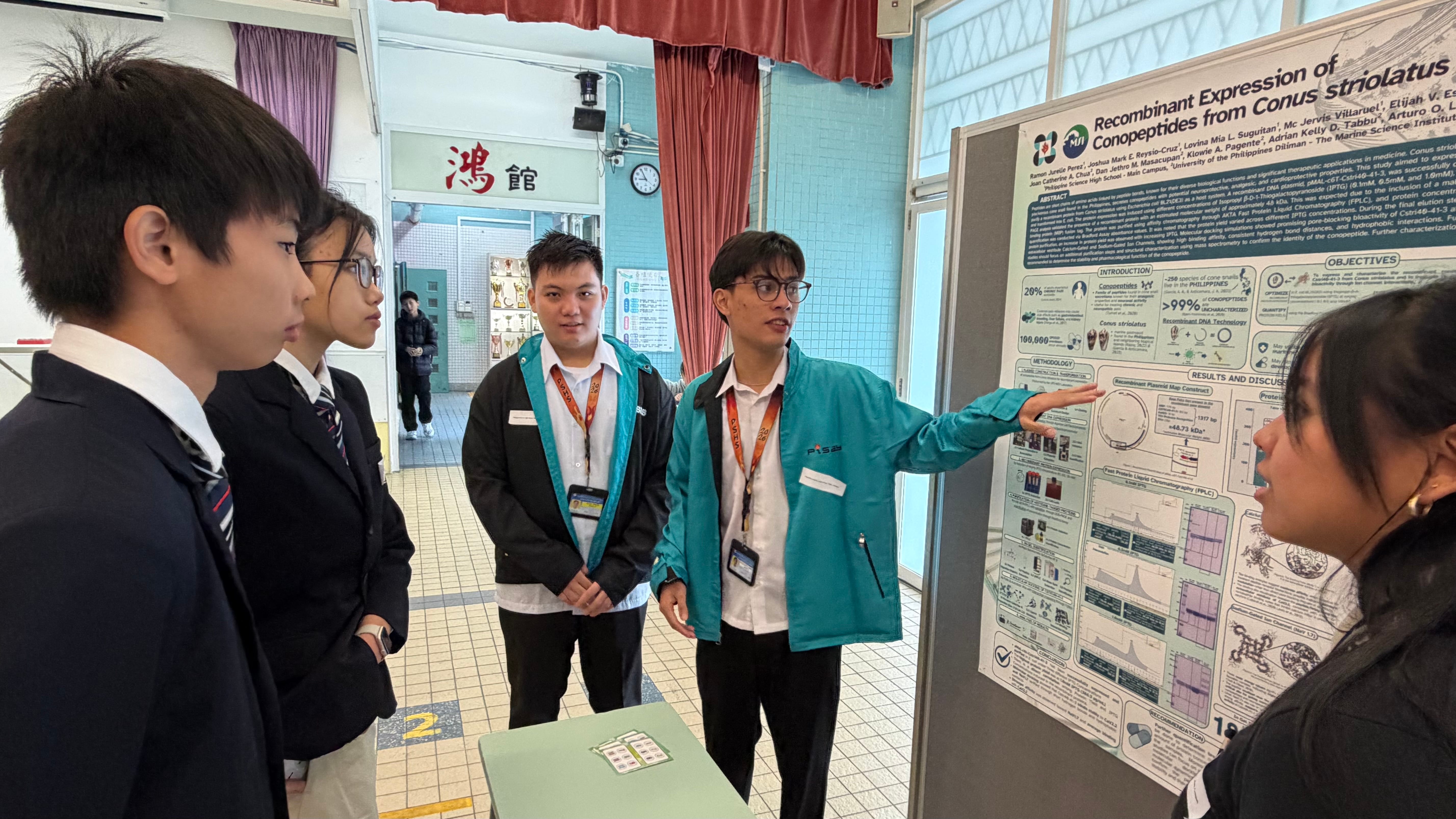 Student in teal jacket points at a research poster while others in uniforms listen in a school hallway.