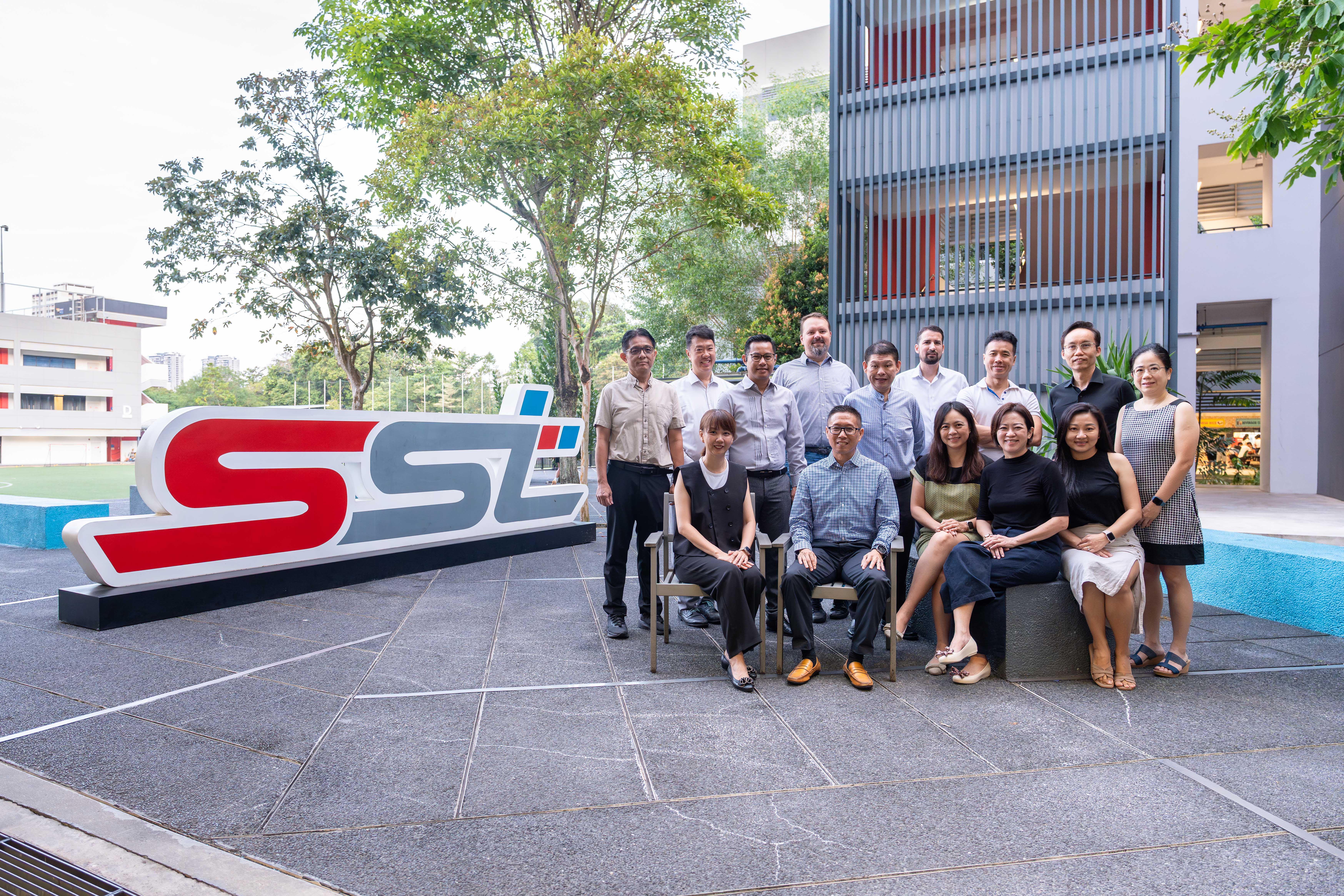 A group of about 14 people posing outdoors near a large red and gray SST sign beside a modern building.