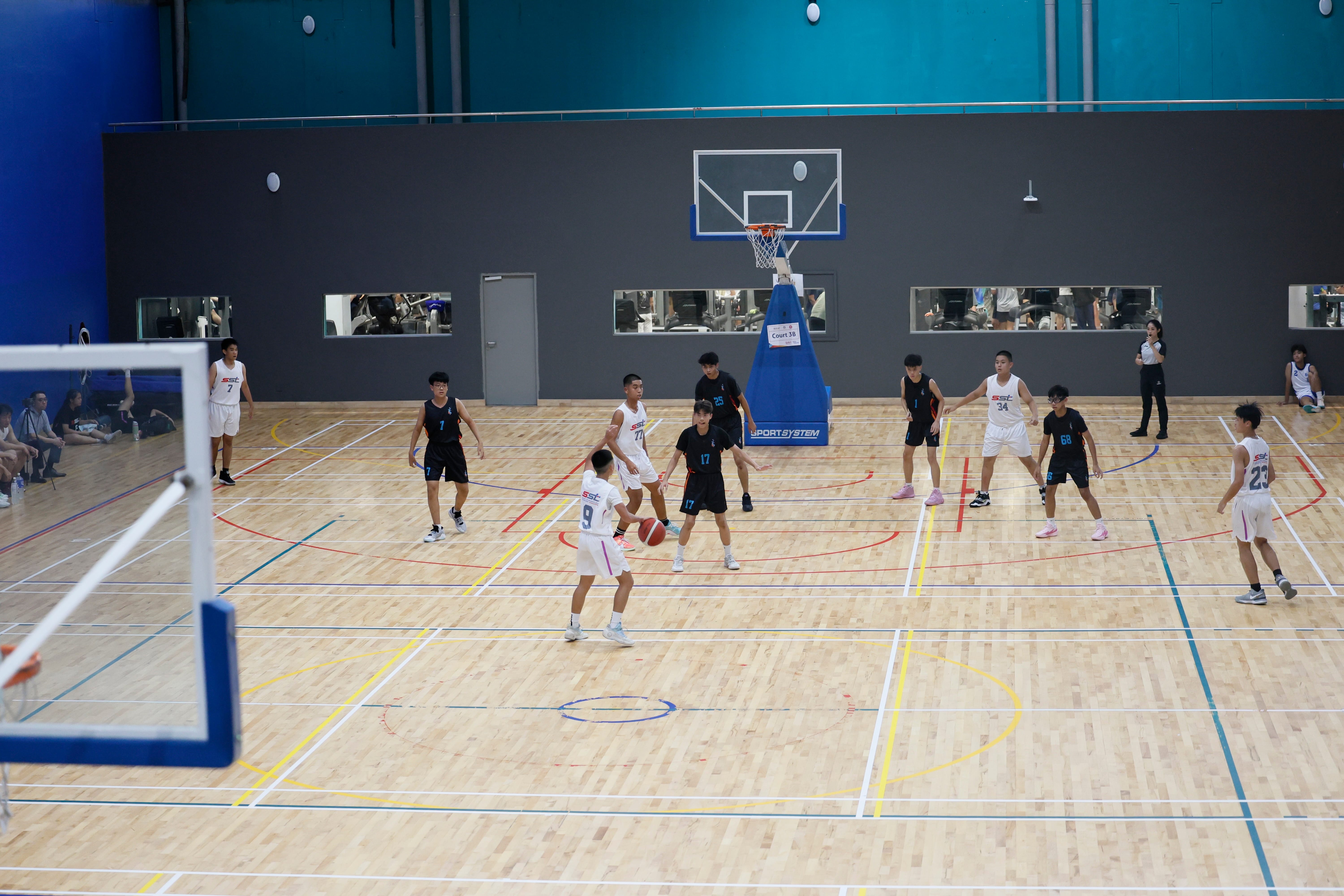 Two teams in white and black uniforms play basketball on an indoor hardwood court with a hoop visible.