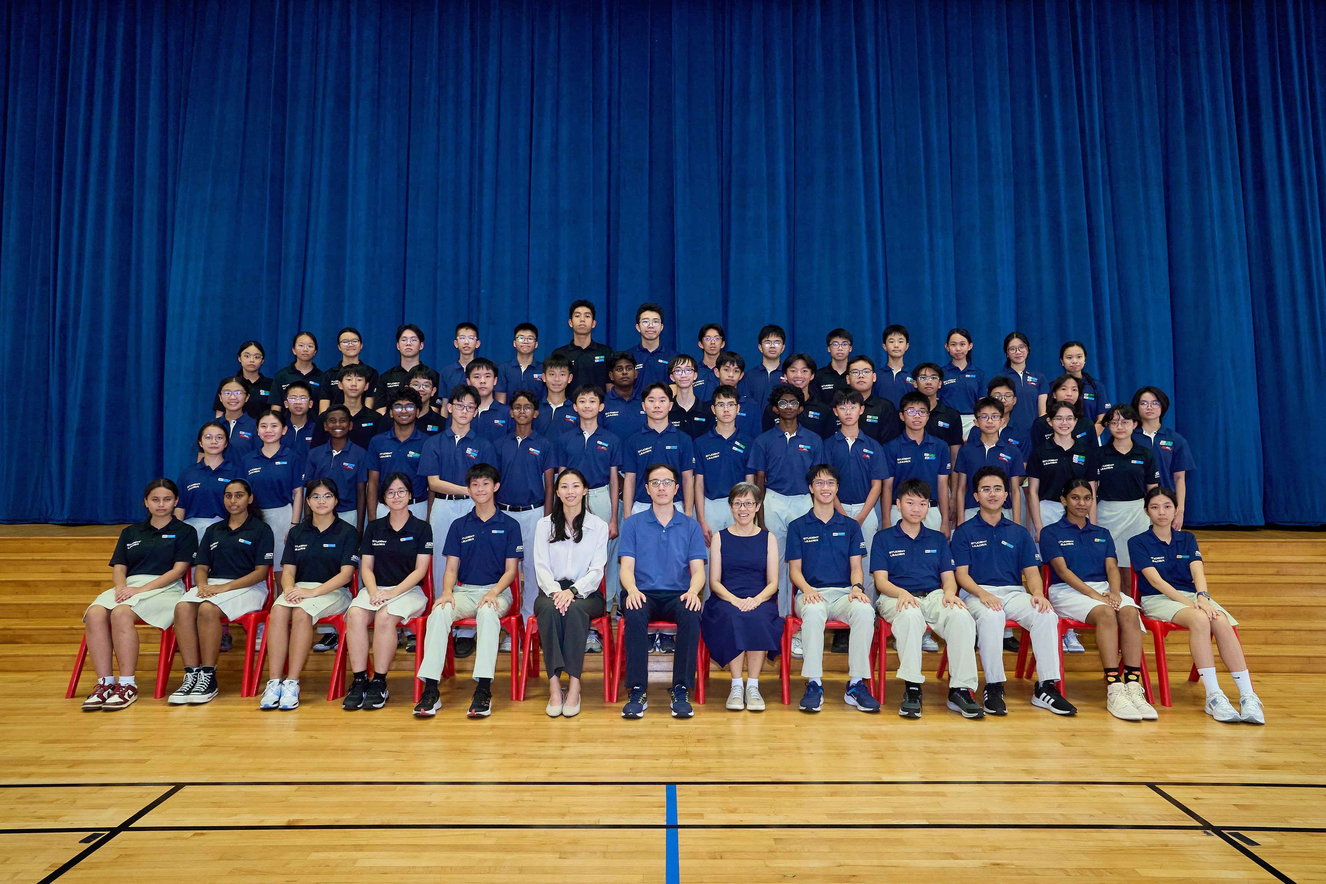 Large group of students and teachers posing in rows on red chairs before a tall blue curtain.