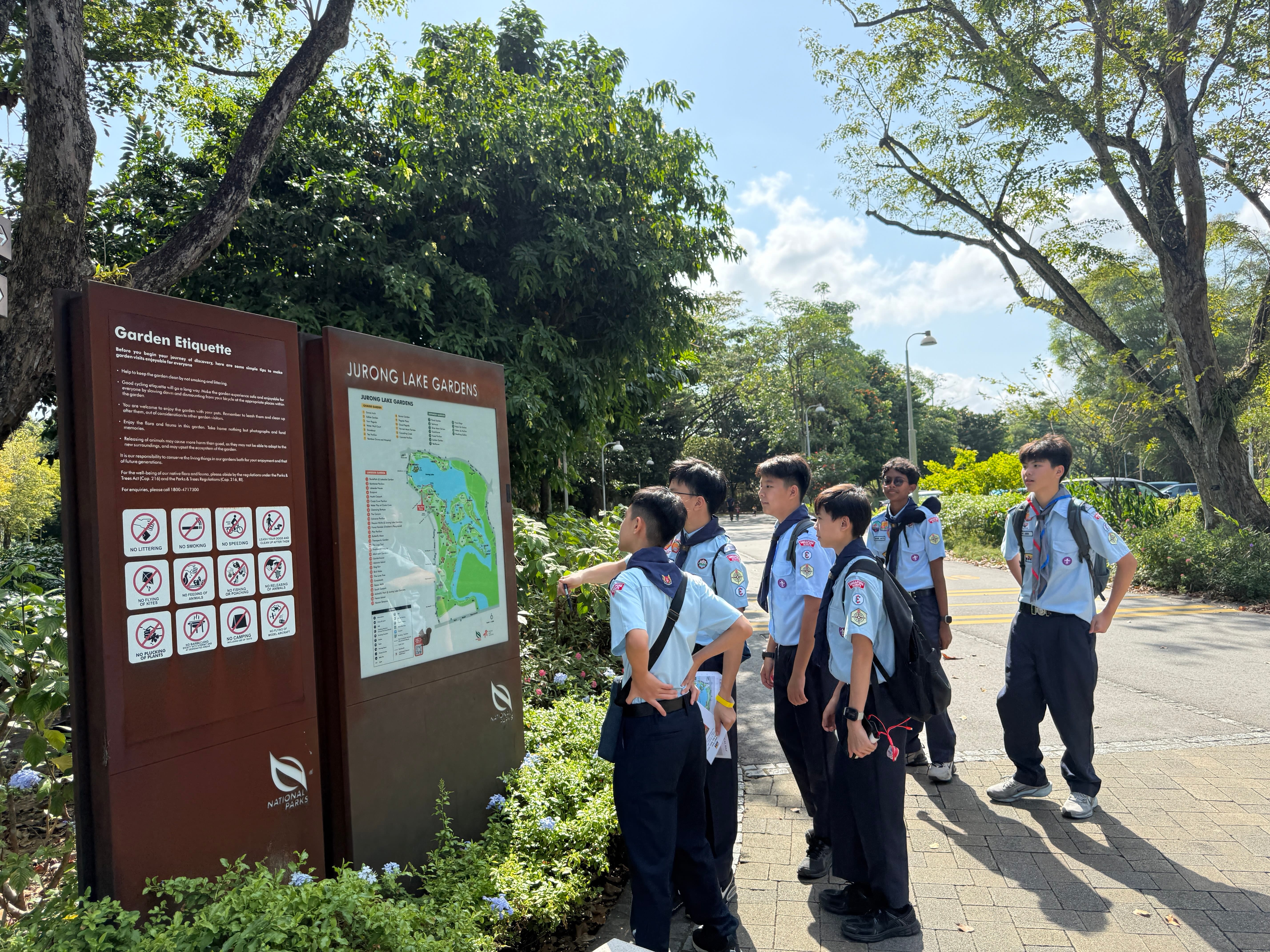 Group of students in uniform read a park map and rules sign beside a sunny, tree-lined path.