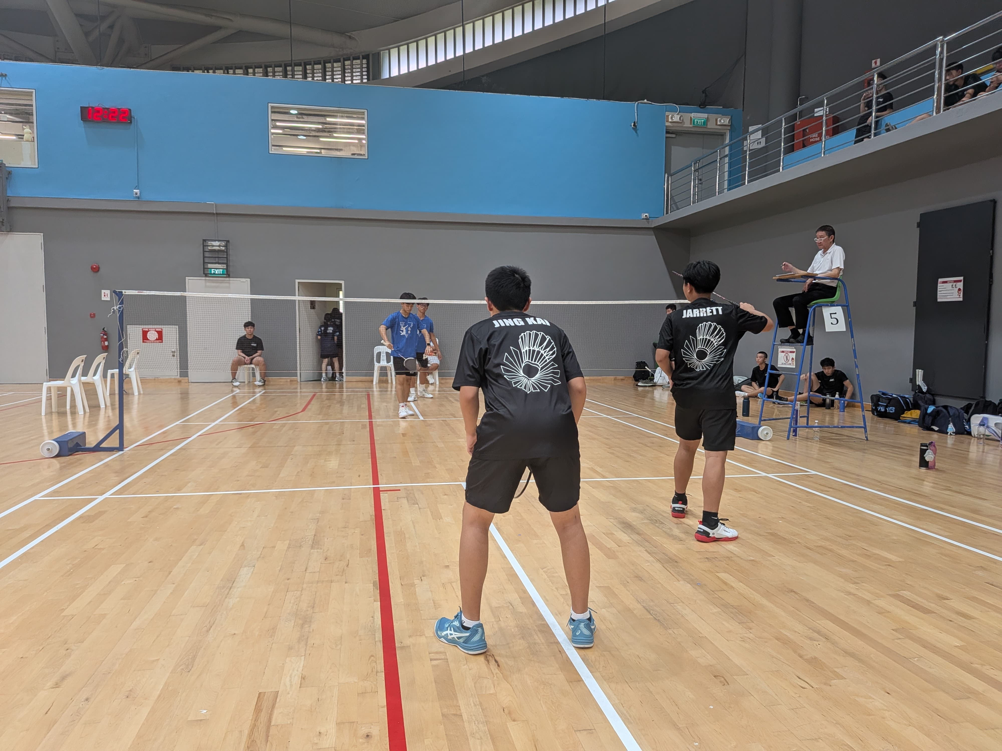 Two players in black shirts named Jing Kai and Jarrett play doubles badminton indoors with a referee on a high chair.