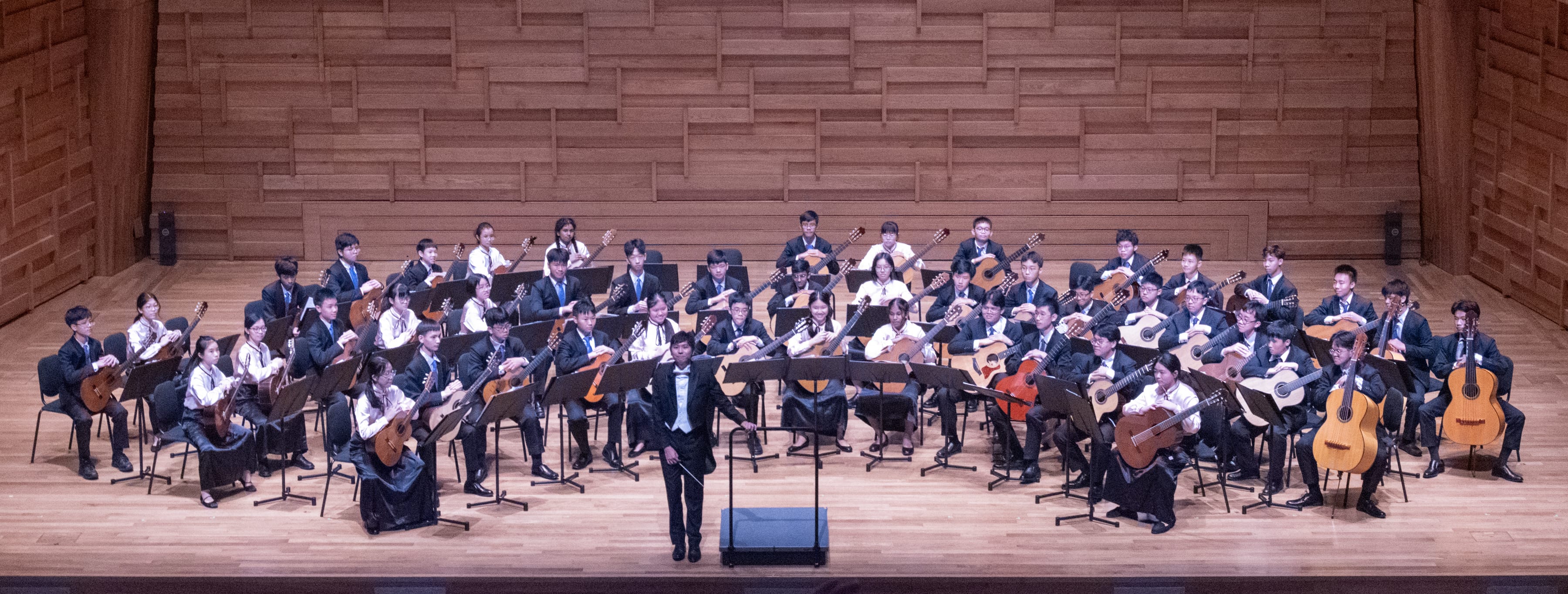 Conductor stands before a seated guitar ensemble on a wooden stage with music stands and many guitars.