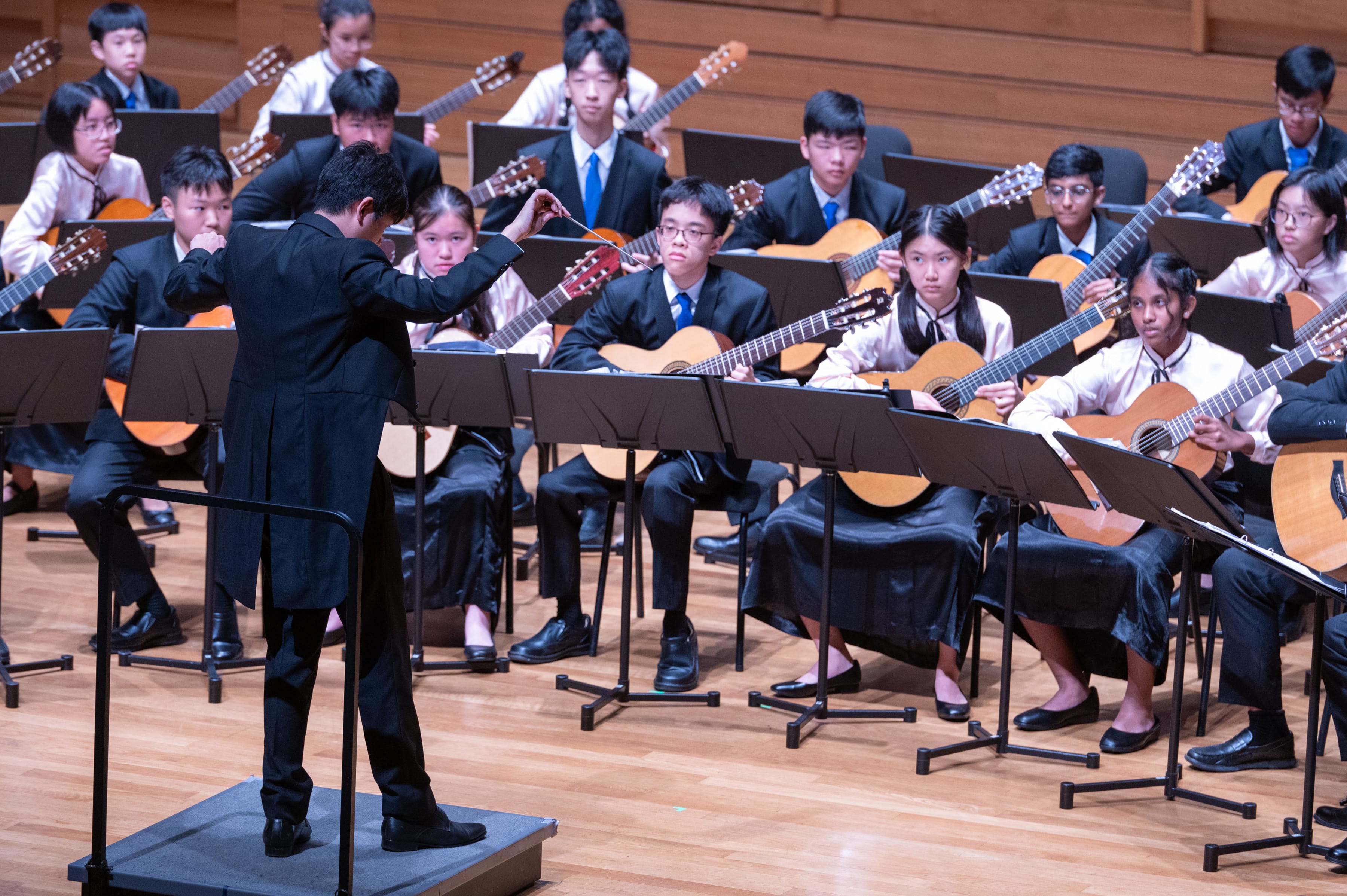 A conductor in black leads a youth guitar ensemble of students in formal attire on a wooden concert stage.
